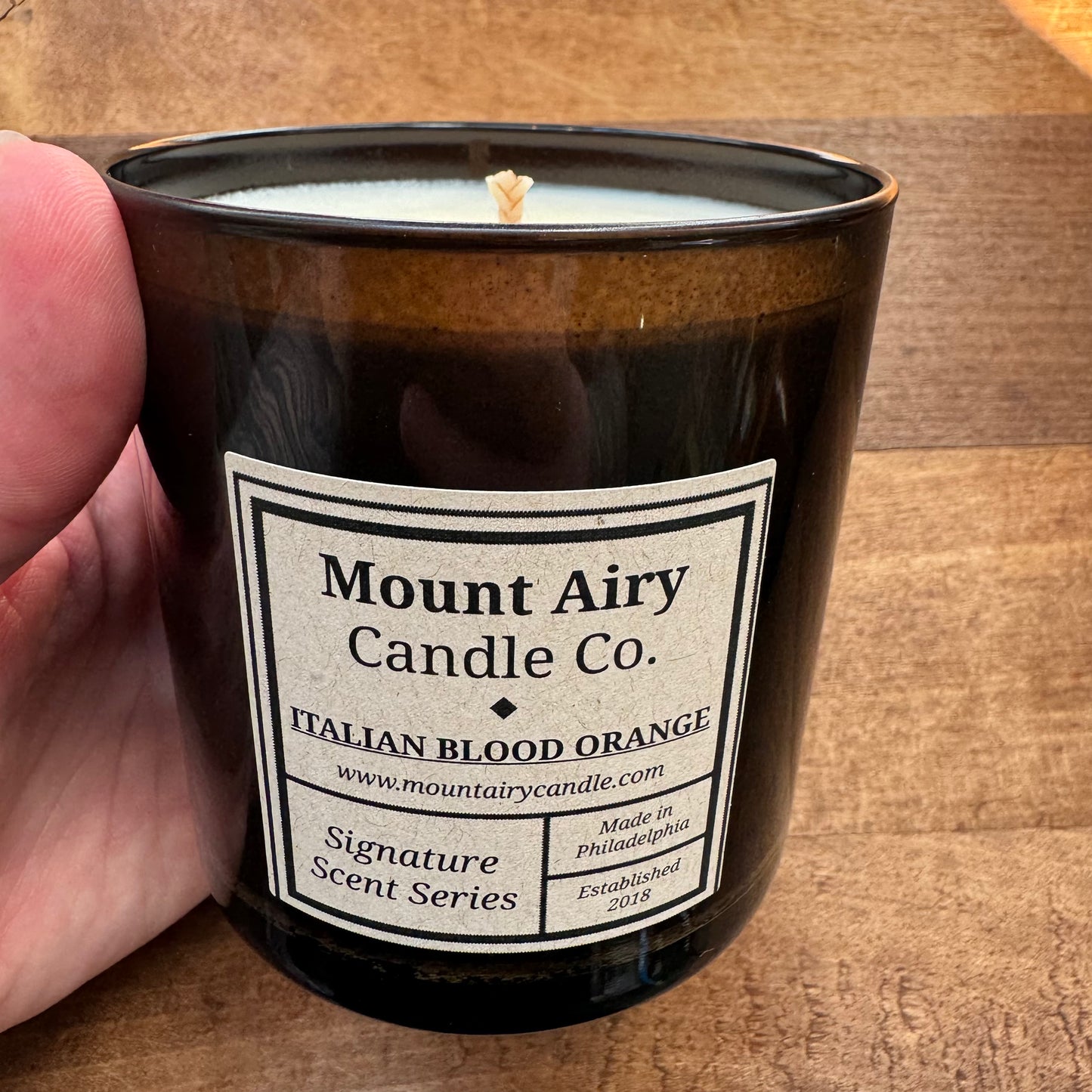 A hand holds a black candle jar labeled "Mount Airy Candle Co., Italian Blood Orange, Signature Scent Series, Est. 2018, Made in Philadelphia." Crafted with hand-poured soy wax for the Mount Airy Candle Co.'s Soy Candles-Glass Tumblers line, this aromatic scent boasts a burn time of 45-55 hours. The jar is set on a wooden surface.