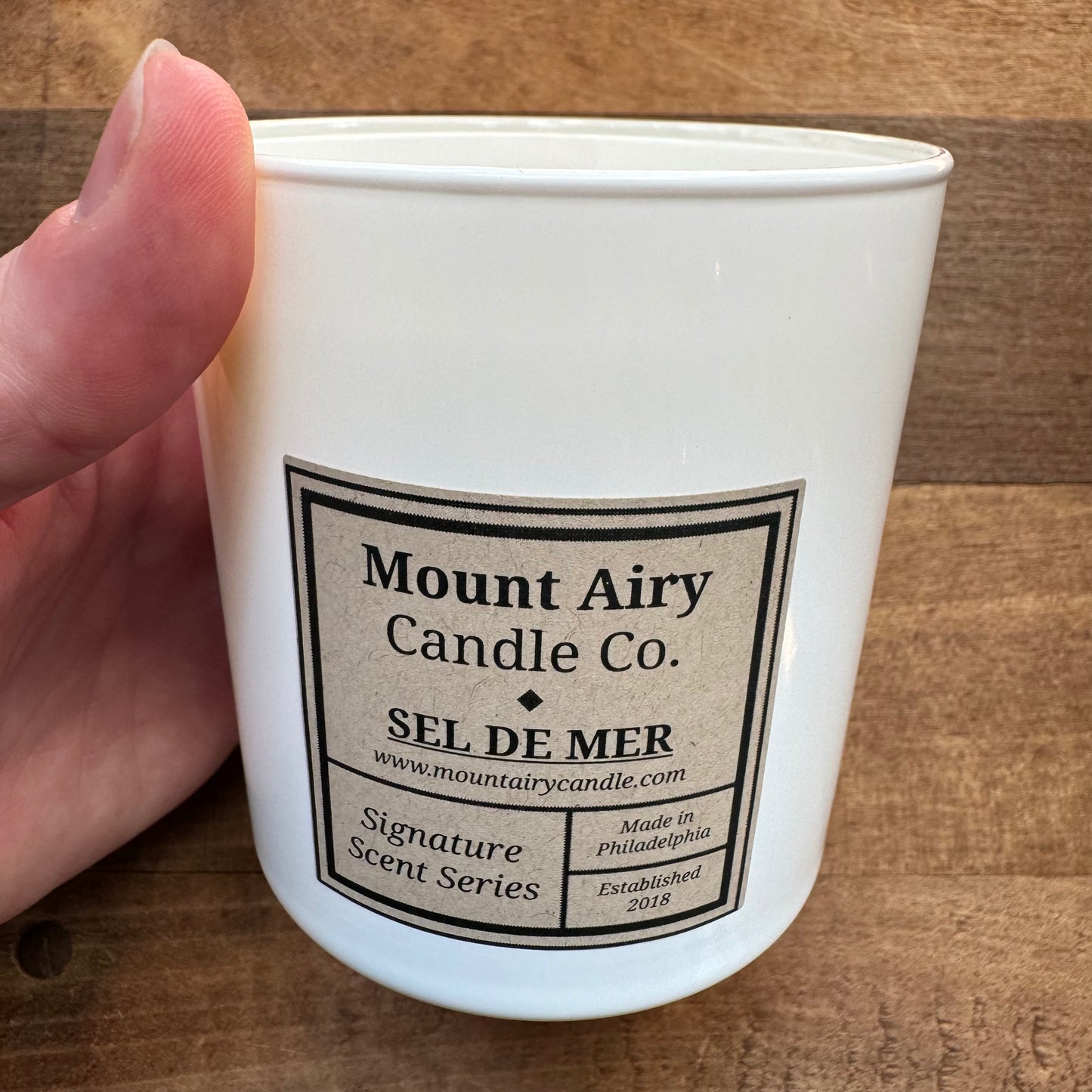 A hand holding a white candle with a label reading "Mount Airy Candle Co. - SEL DE MER - Signature Scent Series - Hand-poured soy wax candles in glass tumblers - Made in Philadelphia - www.mountairycandle.com - Established 2018." Experience aromatic scents with a burn time of 45-55 hours.