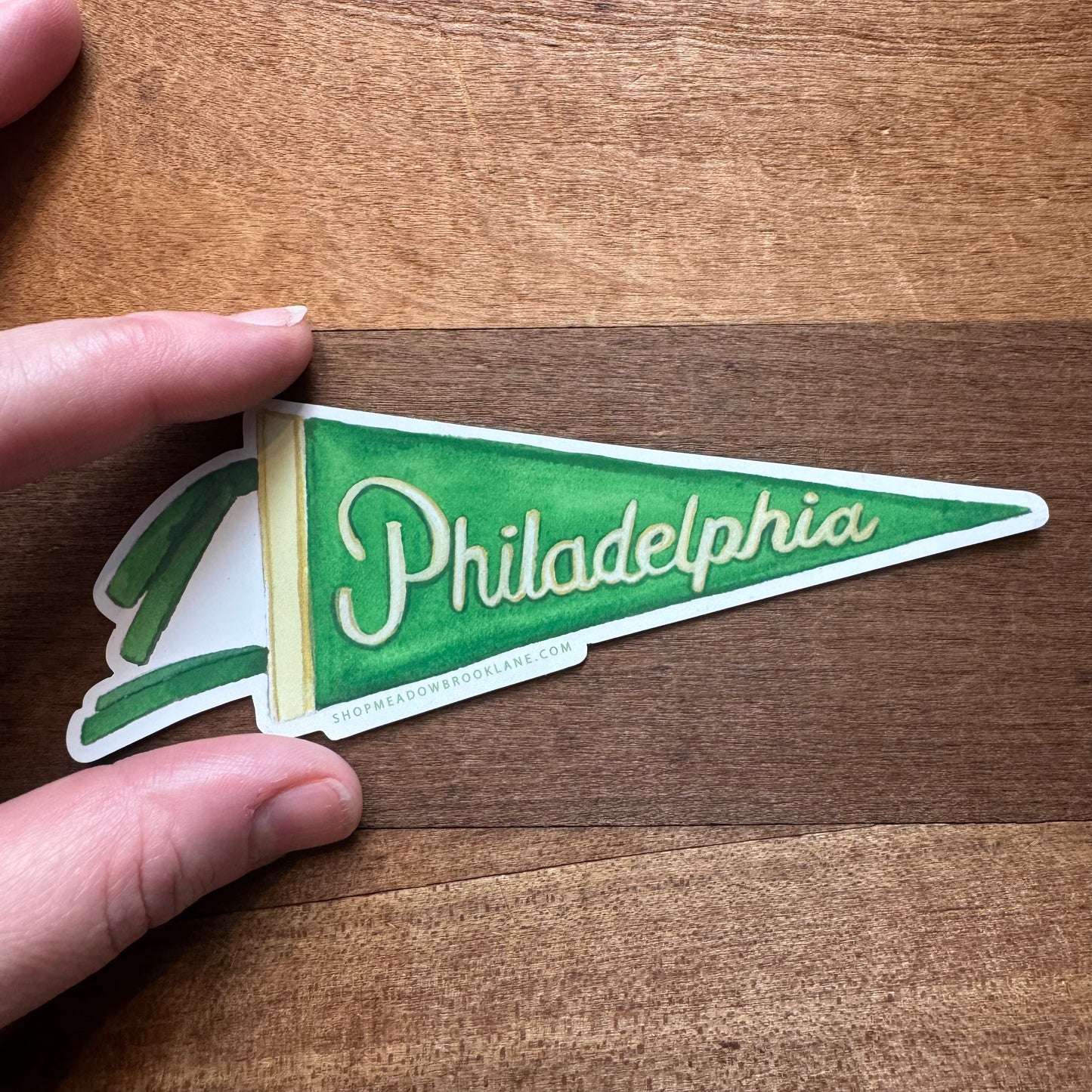 A hand holds a Meadowbrook Lane Philly Vinyl Magnet shaped like a green pennant, adorned with the word "Philadelphia" in crisp white text. The background features a rustic wooden surface.