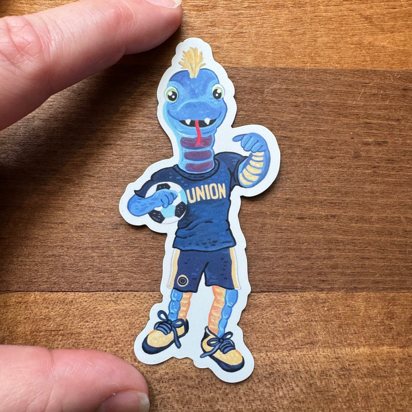 Cartoon sticker of a blue lizard wearing a black "Union" jersey and sneakers, flexing its arms. A person's fingers are holding the sticker on a wooden surface, reminiscent of Gritty's playful spirit. This scene embodies the charm of the Philly Mascot Magnets by Jamie Bendas.