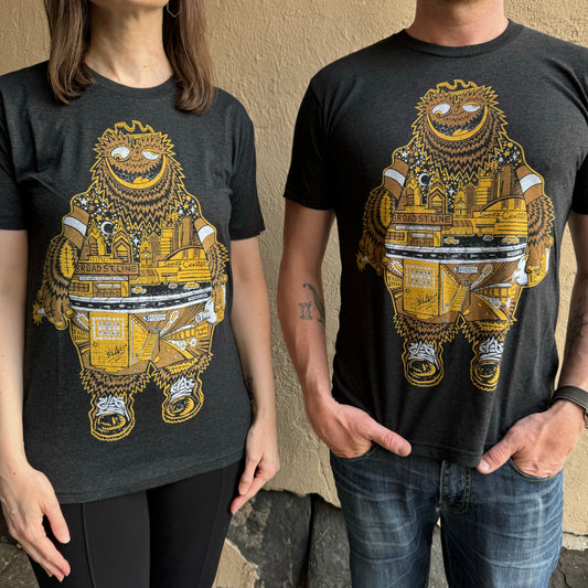 Two people wearing matching heather black unisex Gritty T-Shirts with a colorful, cartoonish character design. The character has a decorated body with various elements, including city buildings and musical symbols from Paul Carpenter.