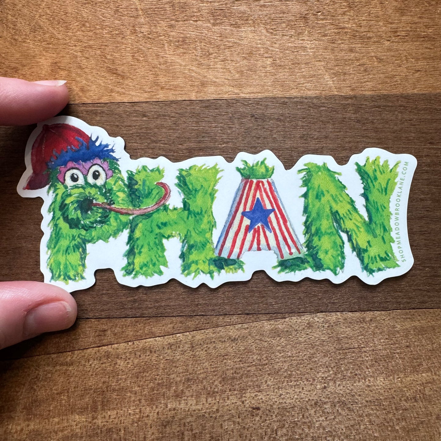 A hand holding a Meadowbrook Lane Philly Vinyl Magnet sticker that spells out "PHAN" in a fuzzy, green, and playful comic-style font, with the "A" being a red and white striped letter featuring a blue star, reminiscent of Philadelphia magnets.