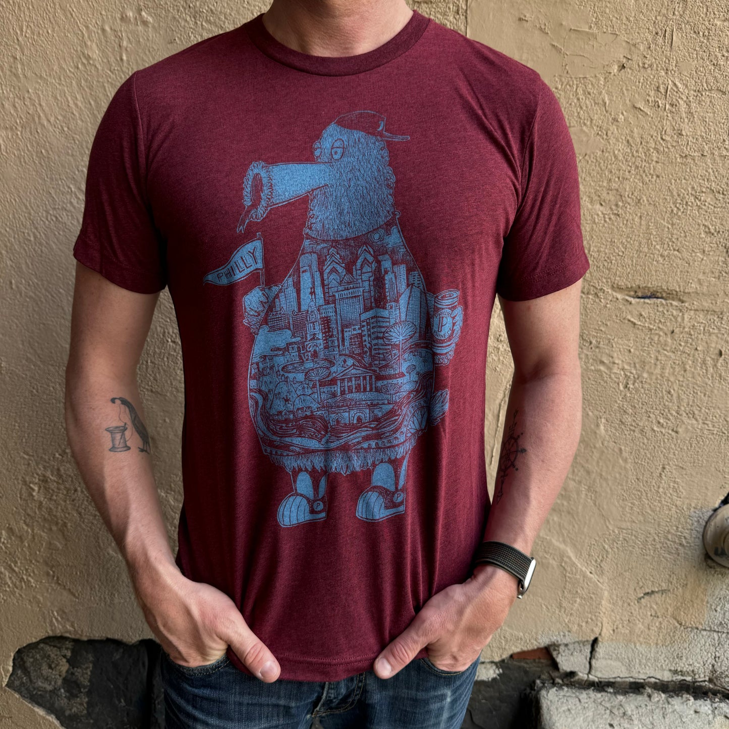 A person in a maroon Phillies Phanatic T-Shirt by Paul Carpenter, standing against a textured beige wall, could easily be mistaken for sporting Phillies mascot attire.