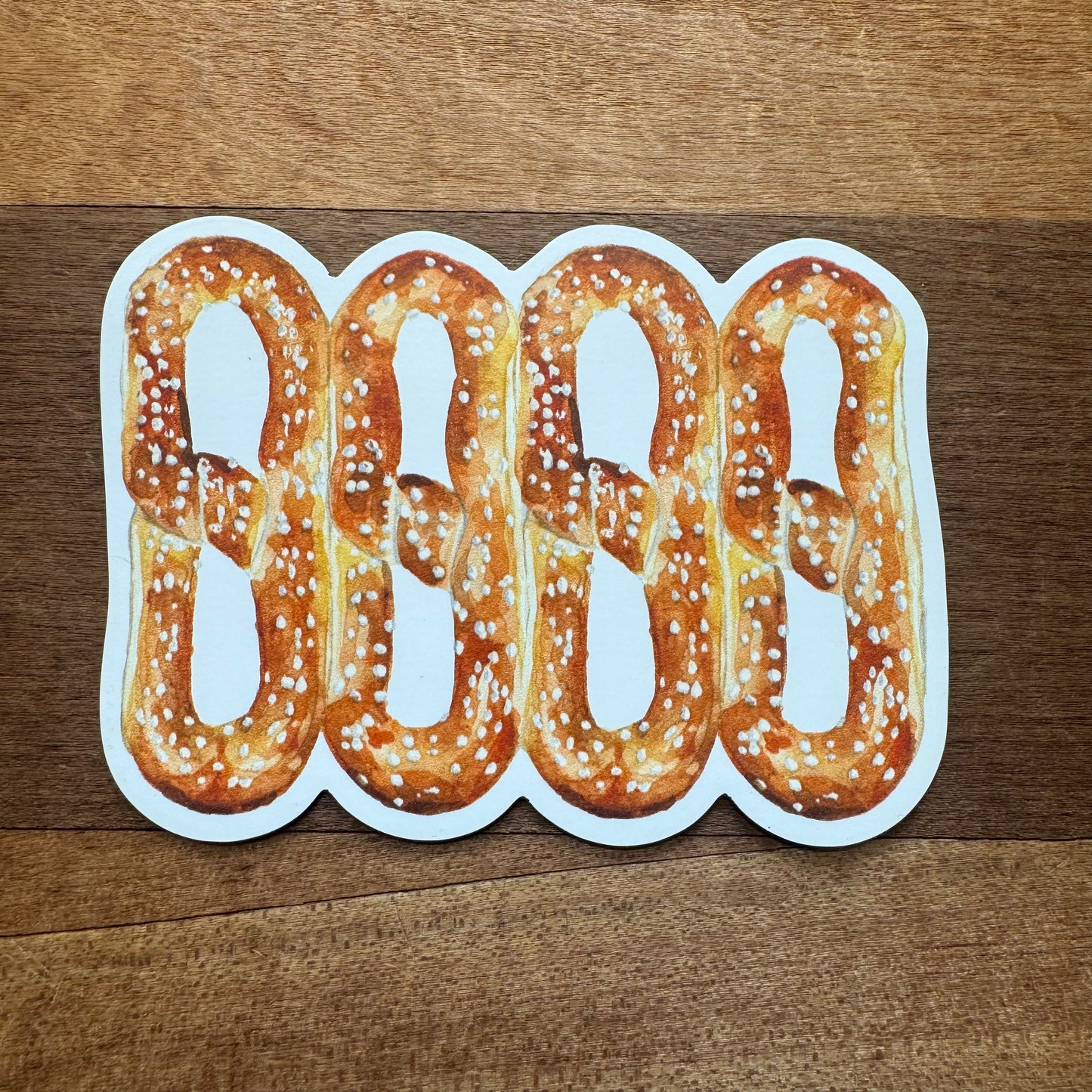 Four illustrated pretzels with salt are arranged in a horizontal line on a wooden surface, perfectly capturing the charming simplicity of Meadowbrook Lane's Philly Vinyl Magnets.