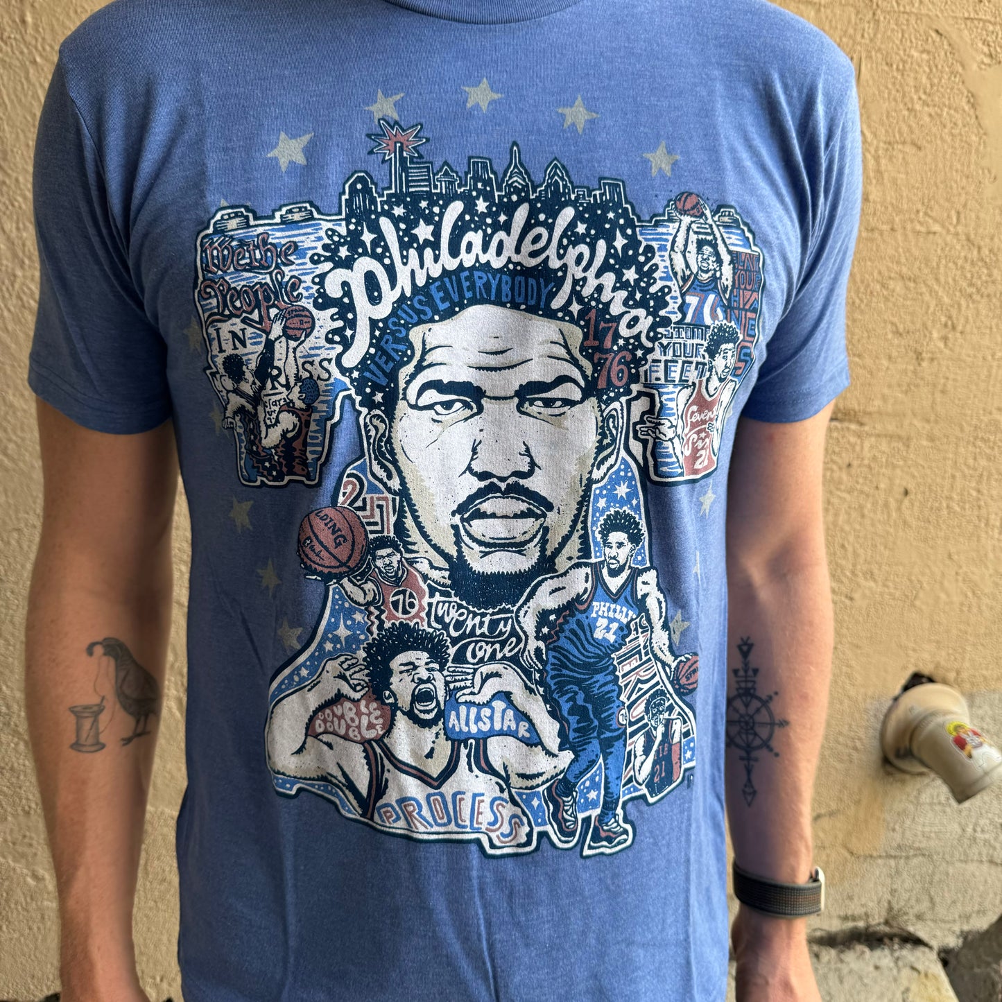 Person wearing a blue unisex Paul Carpenter Sixers Joel Embiid T-Shirt with diverse cartoon illustrations and the word "Philadelphia" prominently displayed among other graphics.