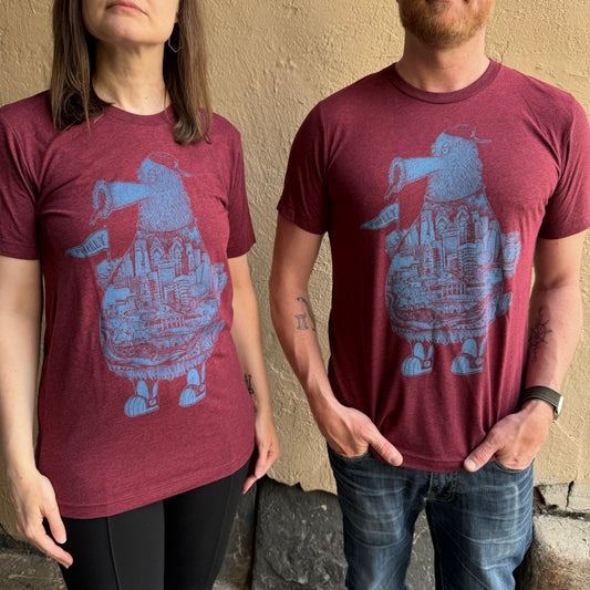 Two people wearing matching maroon Paul Carpenter Phillies Phanatic T-Shirts with a blue graphic of a creature standing on buildings, resembling the Philly skyline. The person on the left has shoulder-length hair, and the person on the right has a short beard.