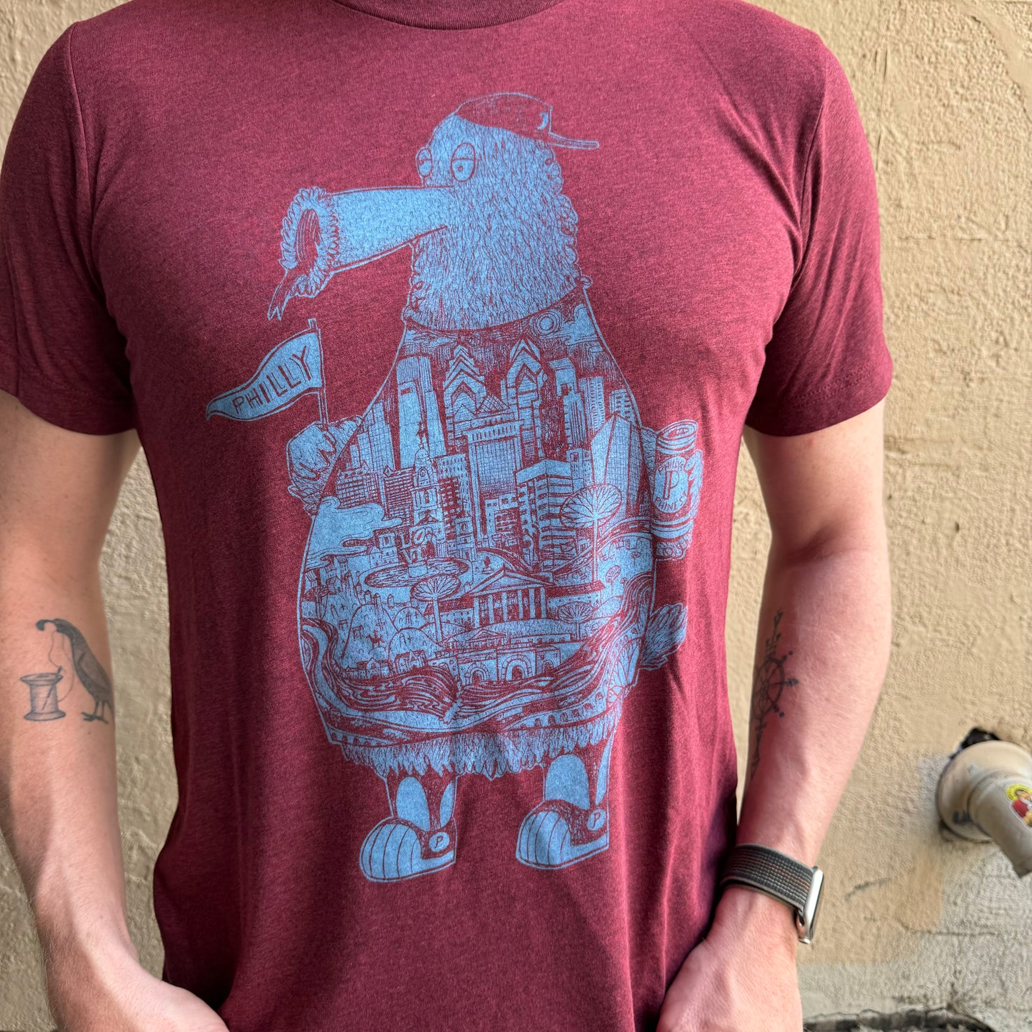 A person wearing a maroon Paul Carpenter Phillies Phanatic T-Shirt featuring a blue illustrated bird holding a Philly flag, with detailed cityscape elements incorporated into the bird's body. The person has tattoos and an Apple Watch.