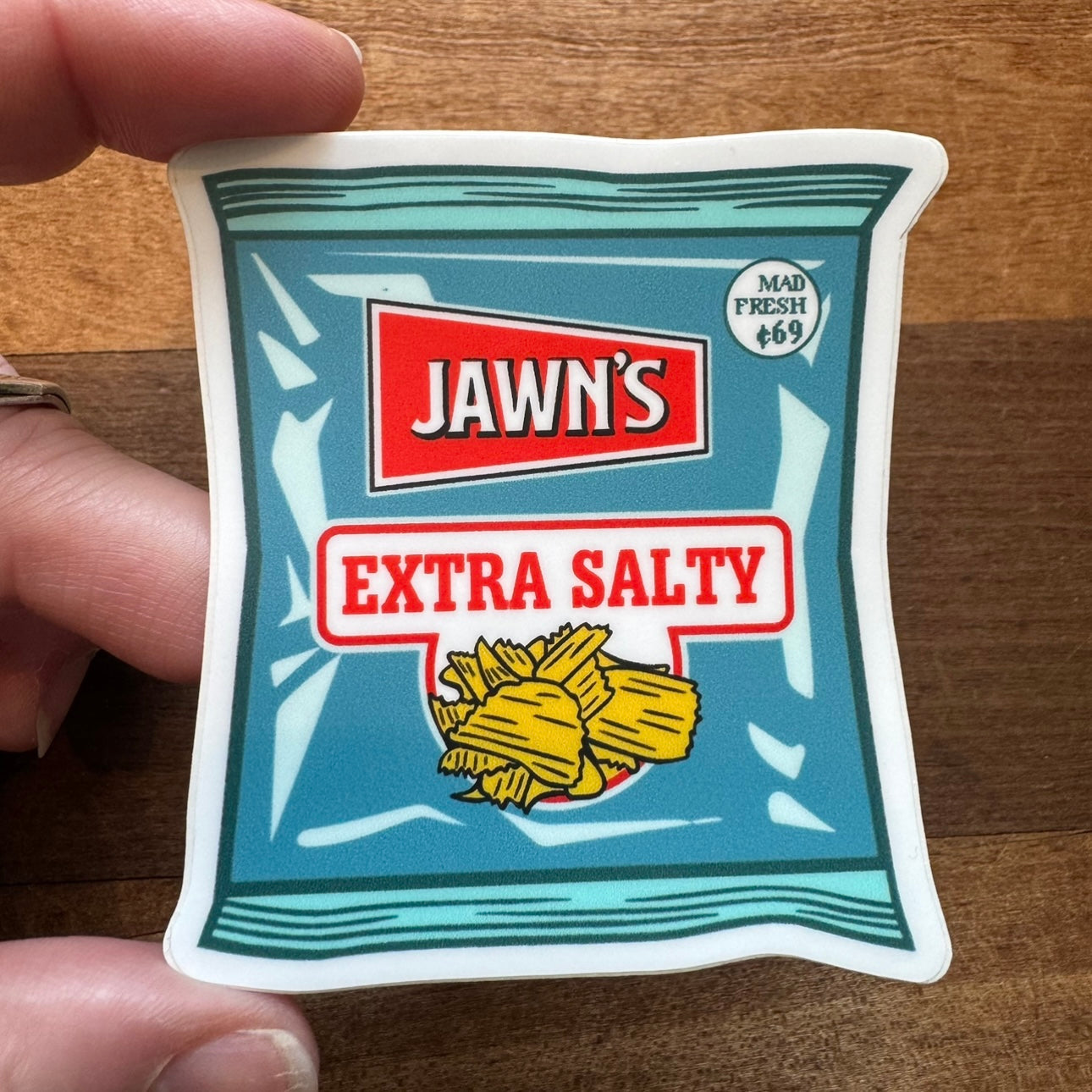 Hand holding a vinyl sticker from the Philly Stickers II collection by South Fellini, shaped like a teal potato chip bag labeled "Jawn's Extra Salty" with an illustration of chips.