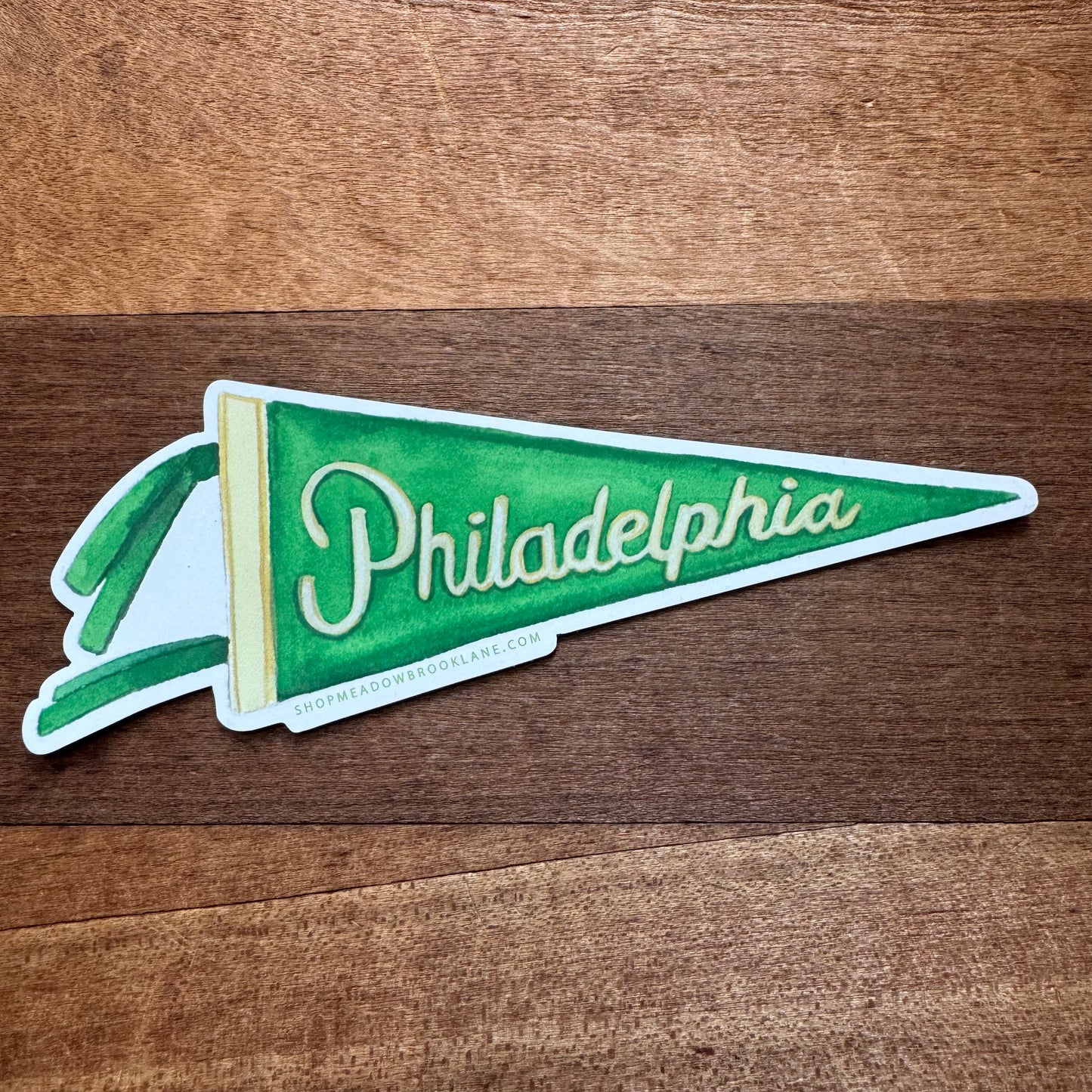 The Philly Vinyl Magnet by Meadowbrook Lane is a green and white pennant-shaped vinyl magnet featuring the word "Philadelphia" in cursive, with a small ribbon accent on the wide end of the pennant. Perfect for Philly-themed decor, it is displayed against a wooden surface background.