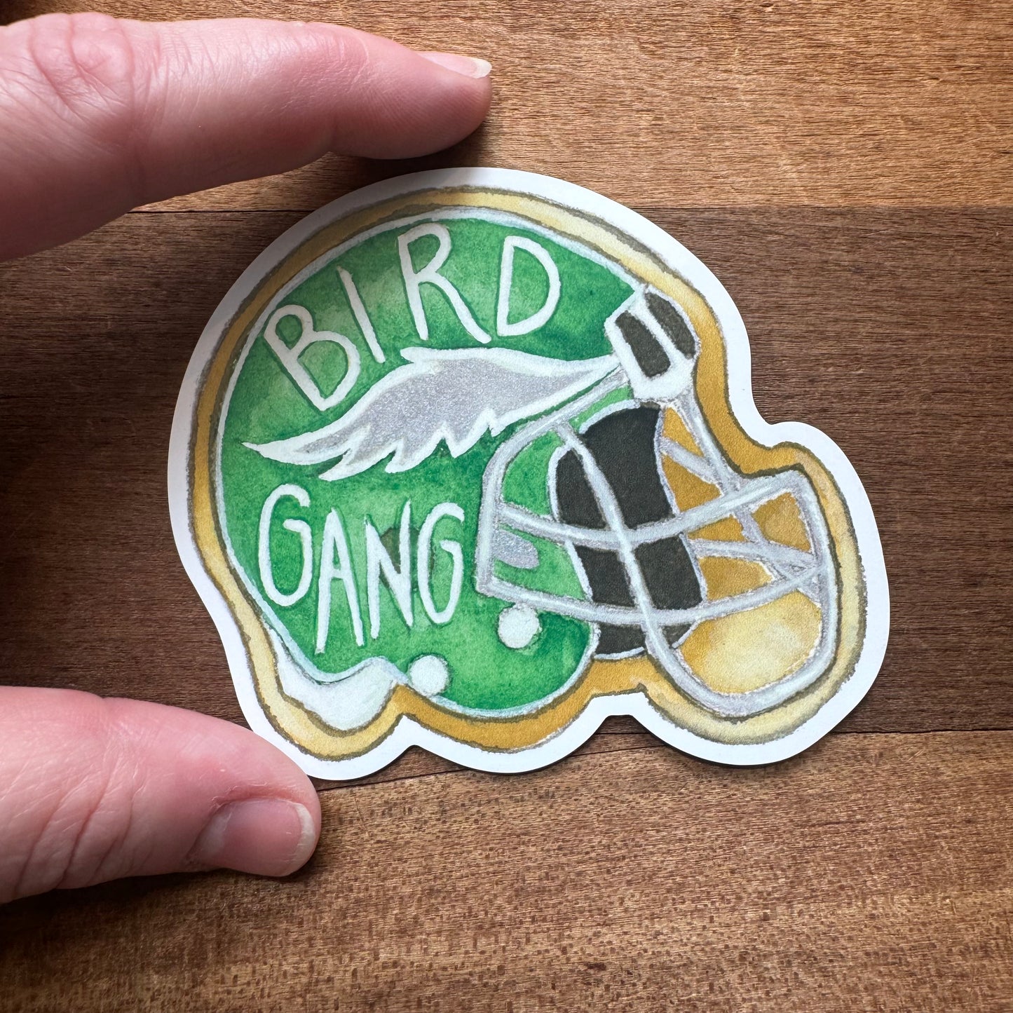 A hand holds a sticker of a green football helmet with the words "BIRD GANG" and a white wing illustration on it. The helmet, complete with a gray facemask, is the perfect addition to any Philly Vinyl Magnets collection by Meadowbrook Lane.