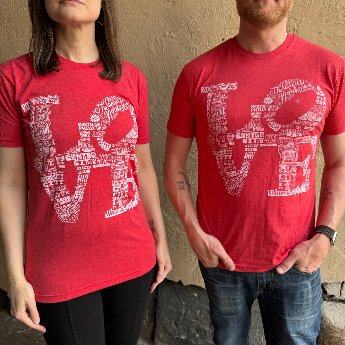 Two people wearing Paul Carpenter's LOVE T-Shirts, each with a "LOVE" design made of various words and locations reminiscent of a Philadelphia neighborhoods t-shirt, stand against a beige wall.