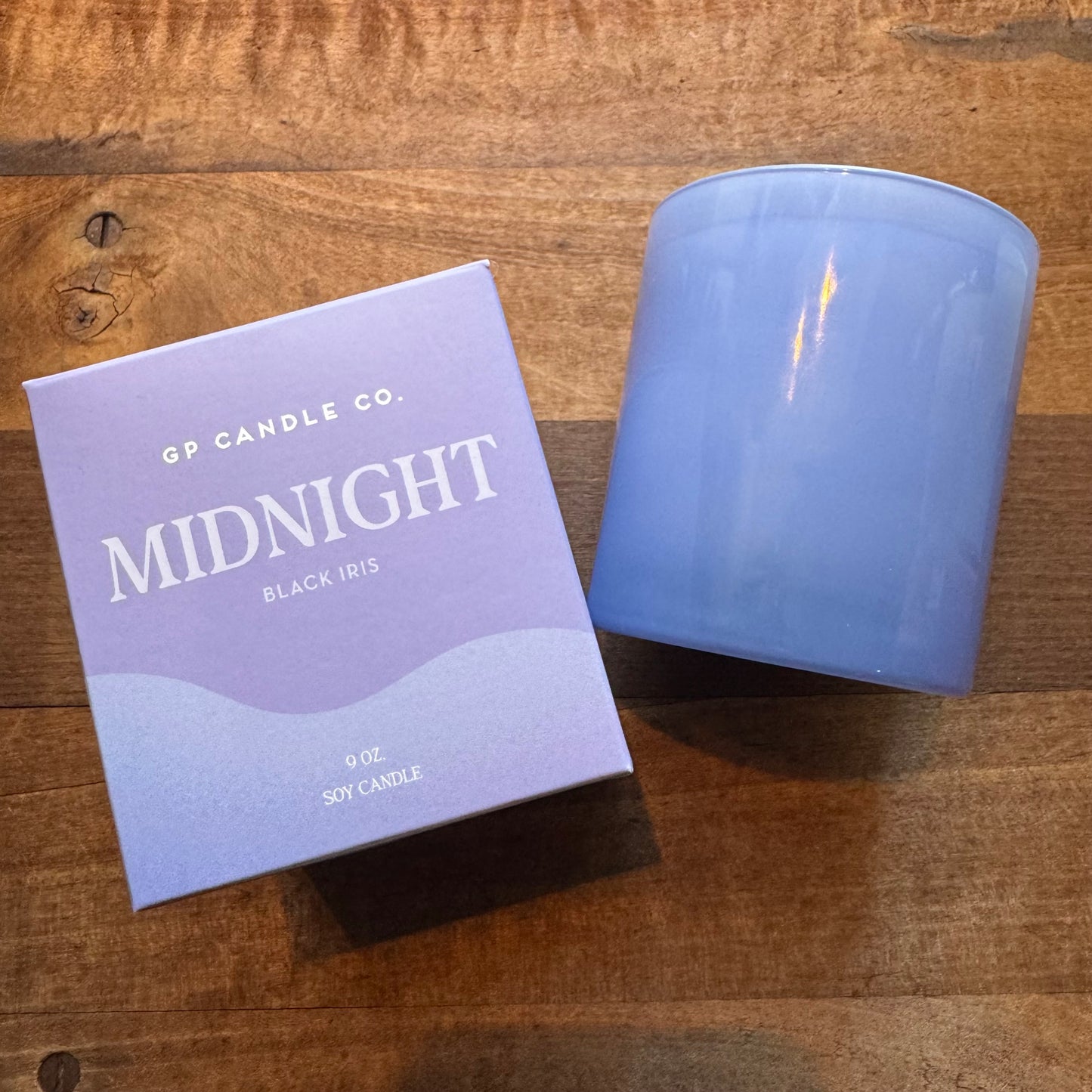 A scented Soy Candles-Hue Collection candle from GP Candle Co. named "midnight" with the scent of black iris next to its packaging box on a wooden surface.