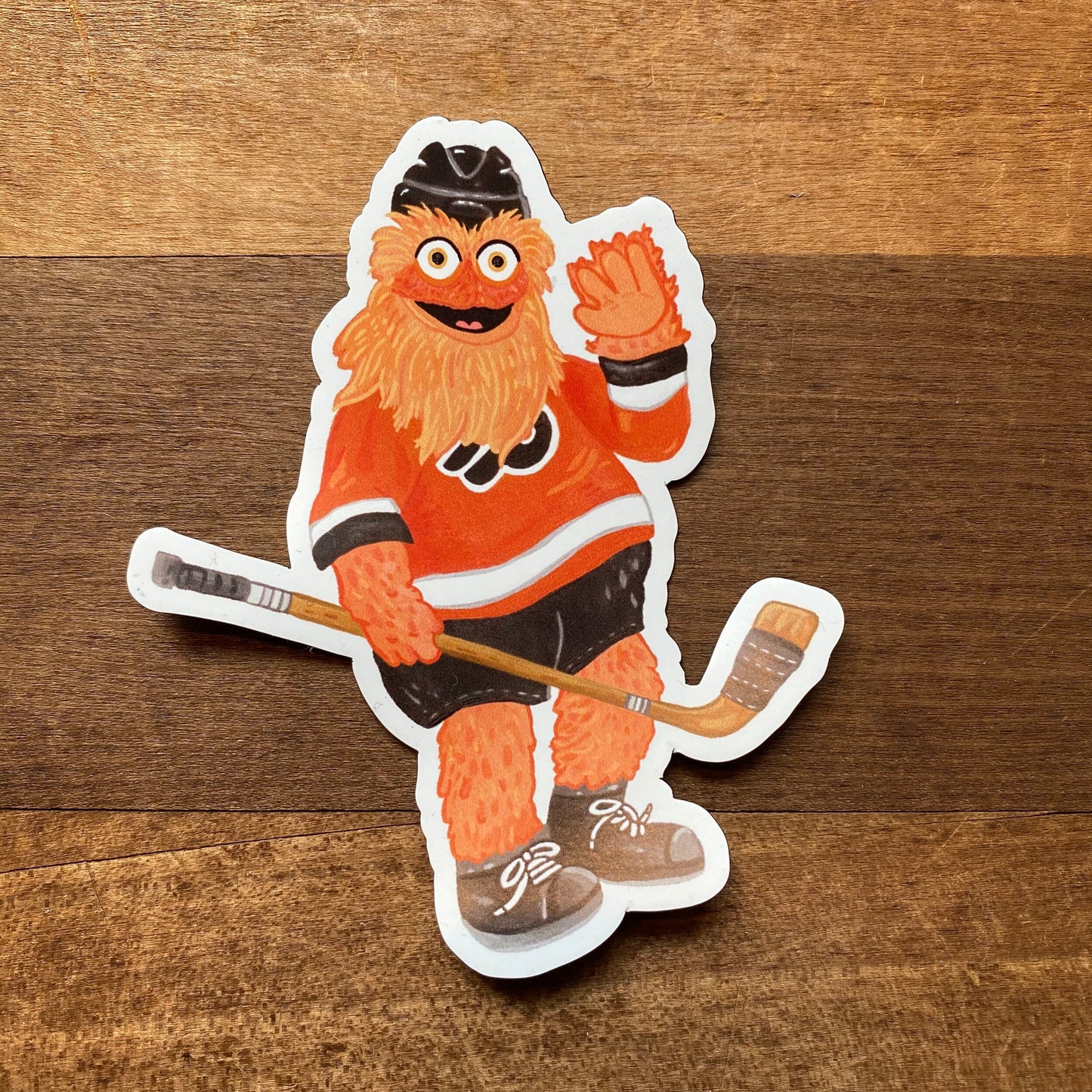 An illustration of Gritty, the mascot for the Philadelphia Flyers hockey team, depicted as a Jamie Bendas Philly Mascot Magnet on a wooden surface.