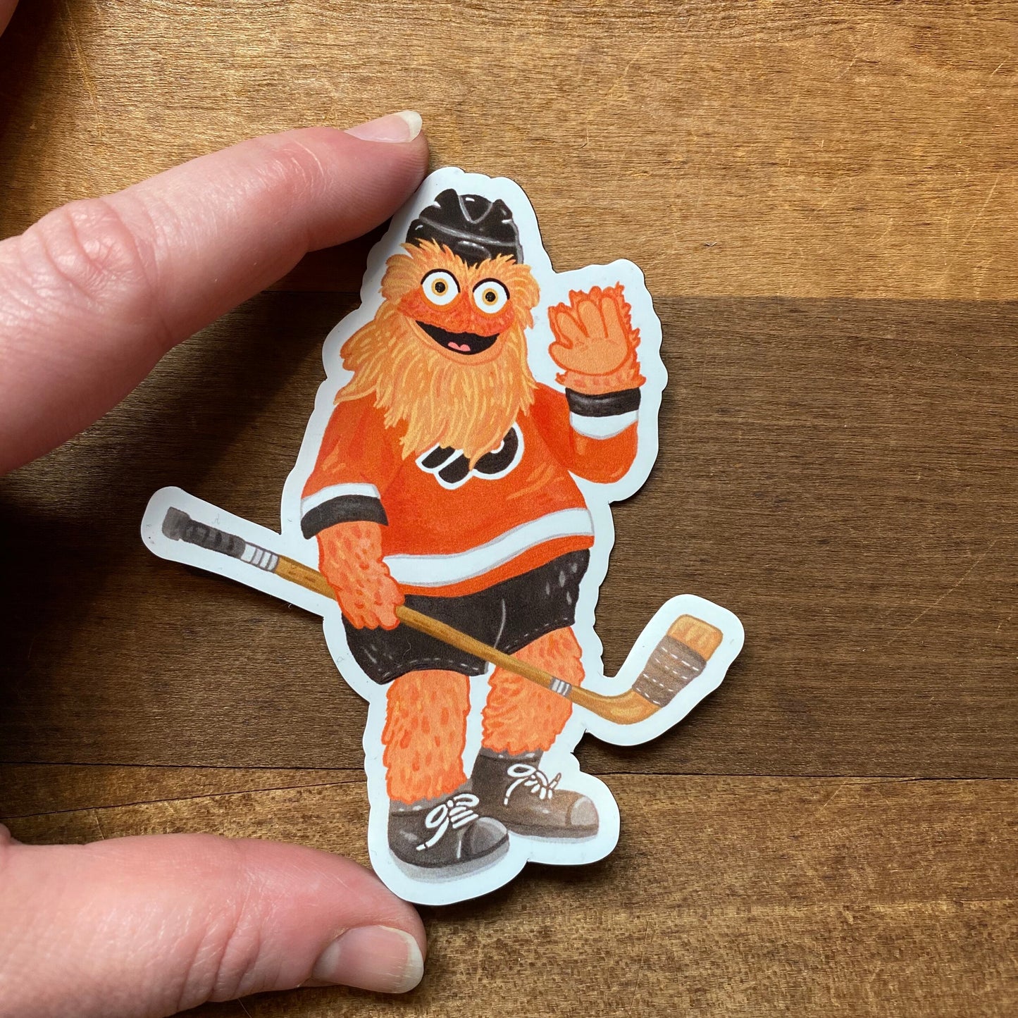 A hand holding a Philly Mascot Magnets sticker of Gritty, a cartoonish hockey mascot, on a wooden surface by Jamie Bendas.