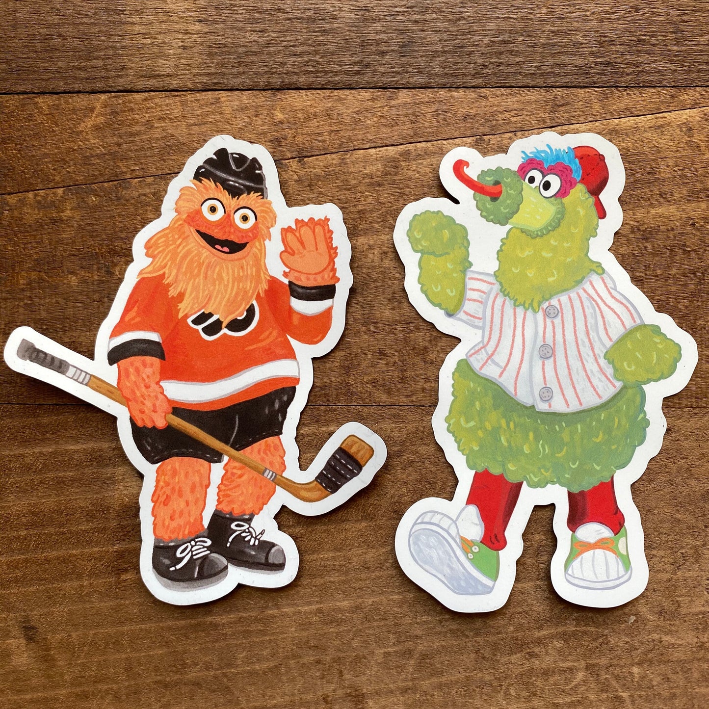 Two Philly Mascot Magnets of cartoonish sports mascots, one dressed as a hockey player and the other in a baseball outfit, placed on a wooden surface by Jamie Bendas.