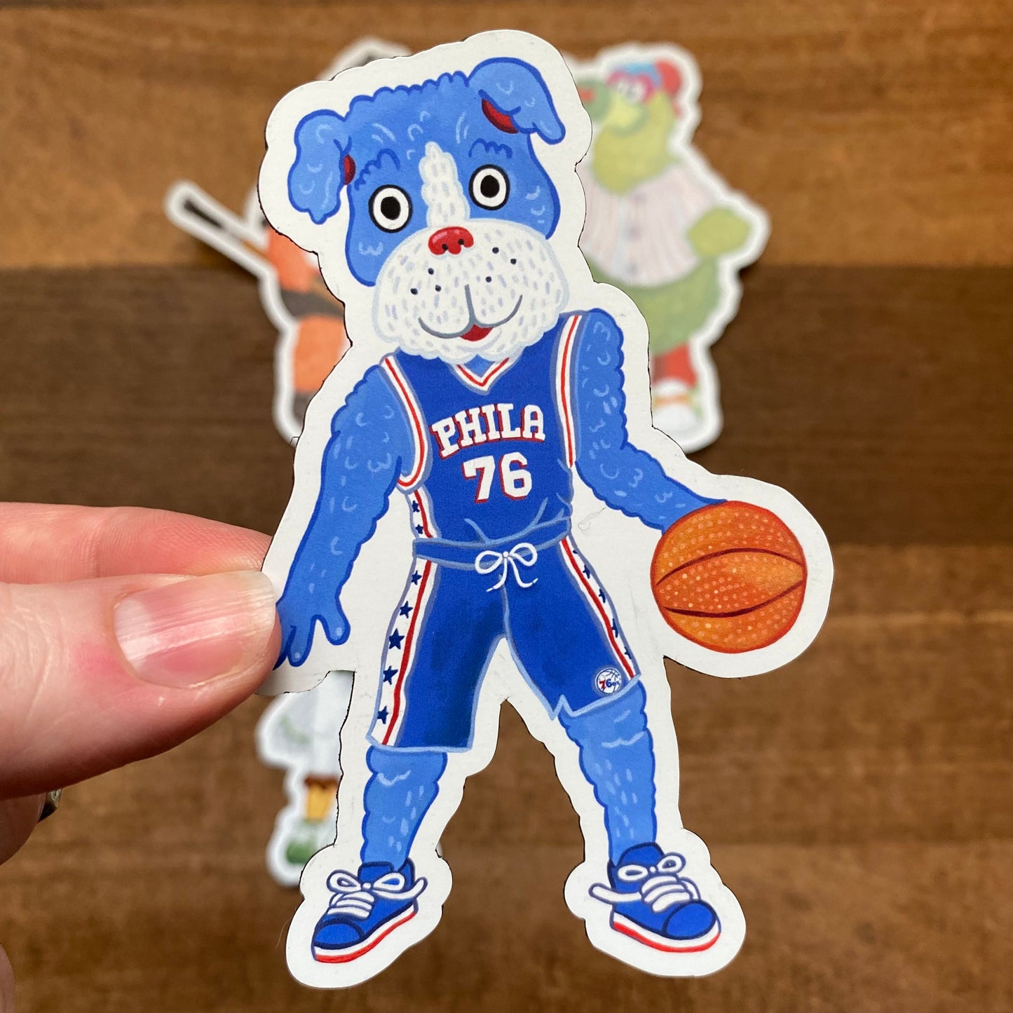 A Philly Mascot Magnet of a cartoon blue dog wearing a basketball uniform with the number 76, holding a basketball, by Jamie Bendas.