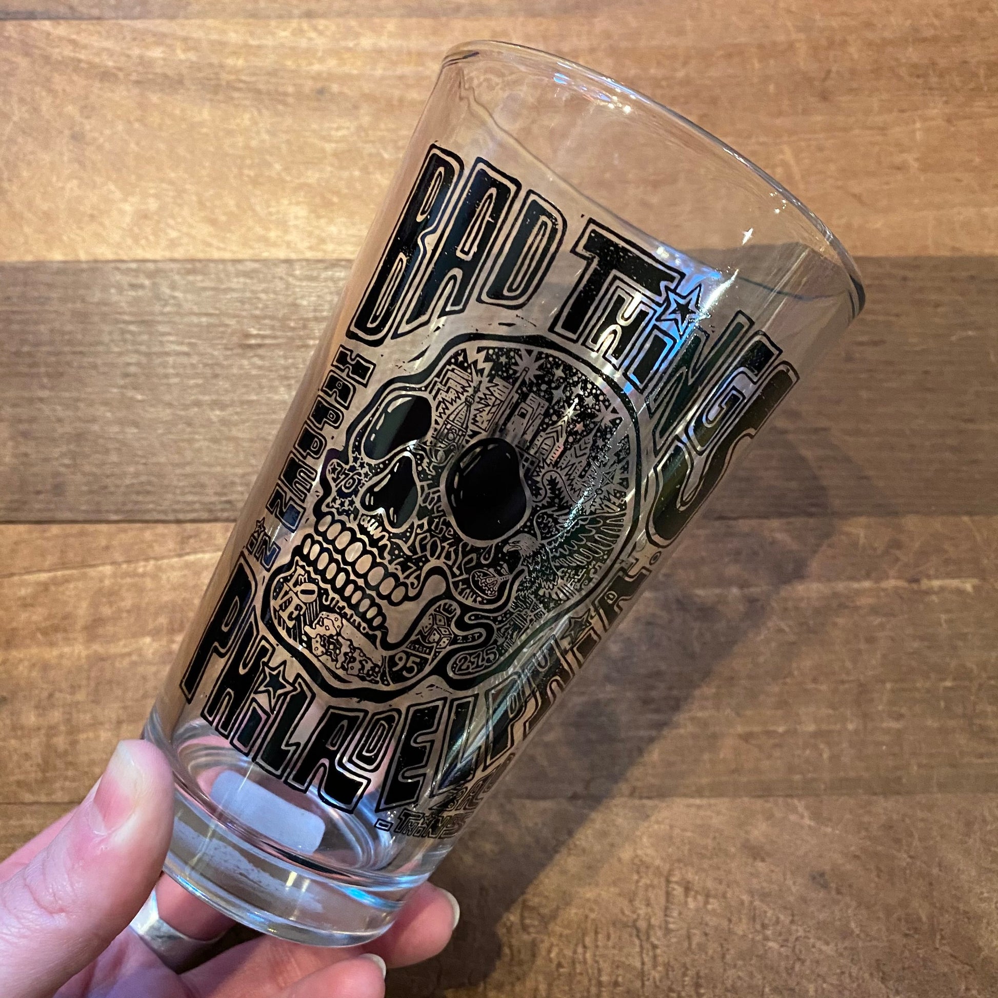 A hand holding an empty Philly Pint Glass featuring a skyline and the text "bad things" printed on it, made by Paul Carpenter.