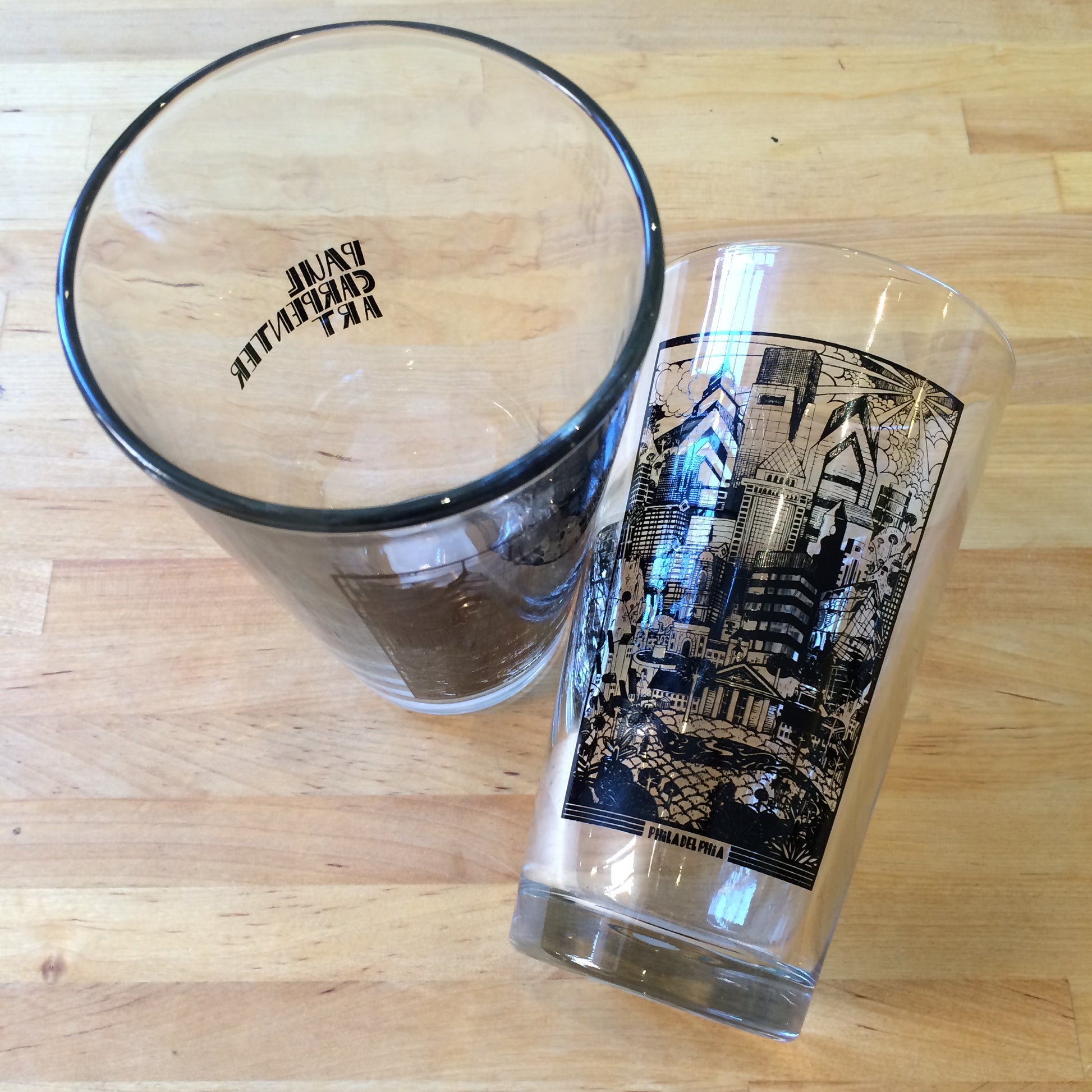 Two empty Philly Pint Glasses on a wooden surface, one upright with a Philadelphia-themed skyline design and one tipped over. Brand: Paul Carpenter.