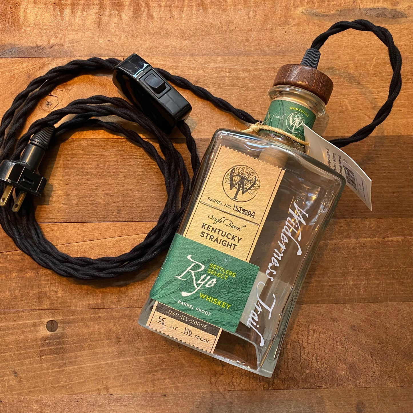 A Salvage & Craft bottle of Kentucky straight rye whiskey transformed into a Bottle Pendant Lamp with an Edison bulb, hung by a decorative lamp cord over a wooden table.