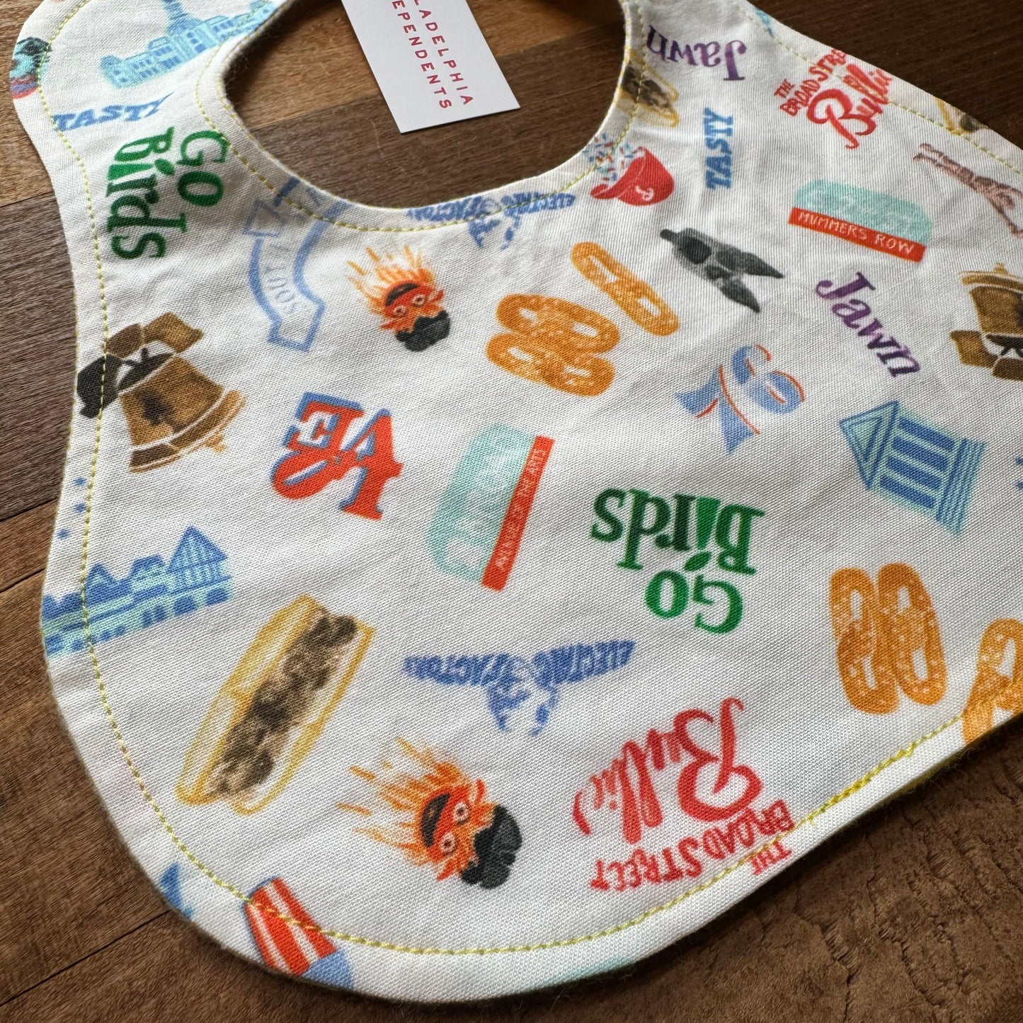 A patterned cotton Philly Print Baby Bib with assorted Sew Much Cooler food and beverage brand logos displayed on a wooden surface.
