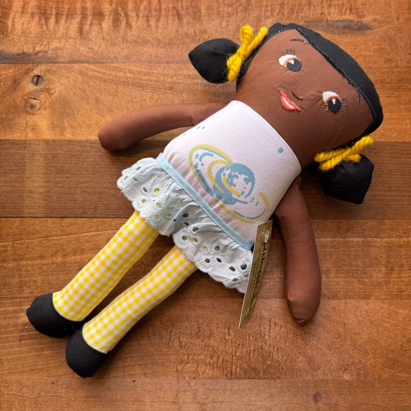 A one-of-a-kind Useful & Beautiful Handmade fabric doll with dark skin, pigtails, and a white and blue dress, lying on a wooden surface.