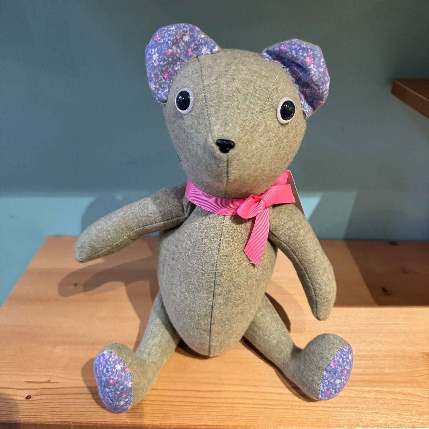 A Useful & Beautiful Handmade stuffed teddy bear with grey fabric, floral ear accents, and a pink bowtie, sitting on a wooden shelf, perfect as a baby toddler gift.
