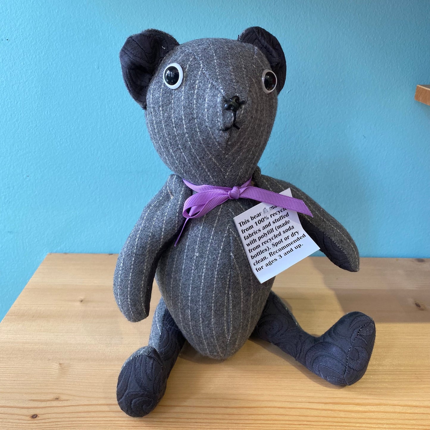 A handmade upcycled Useful & Beautiful Handmade stuffed teddy bear with black eyes and a purple bow, sitting on a wooden table against a teal background. A tag with text is attached to its neck.