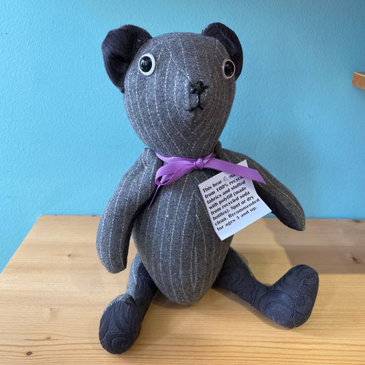 A handmade upcycled Useful & Beautiful Handmade stuffed teddy bear with black eyes and a purple bow, sitting on a wooden table against a teal background. A tag with text is attached to its neck.