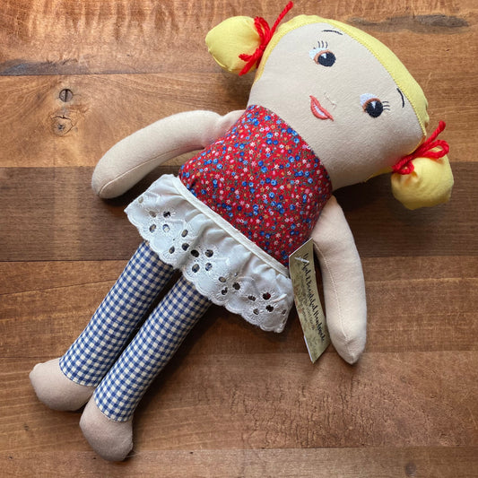 A unique Useful & Beautiful Handmade cloth doll with yellow hair, wearing a patterned red dress and checkered pants, lies on a wooden surface.
