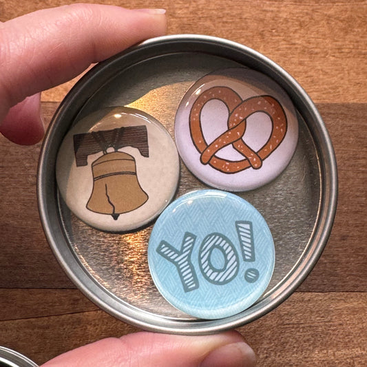 A tin containing three graphic pins: a coffee cup, an exit343design Philly Pretzel magnet, and one with the word