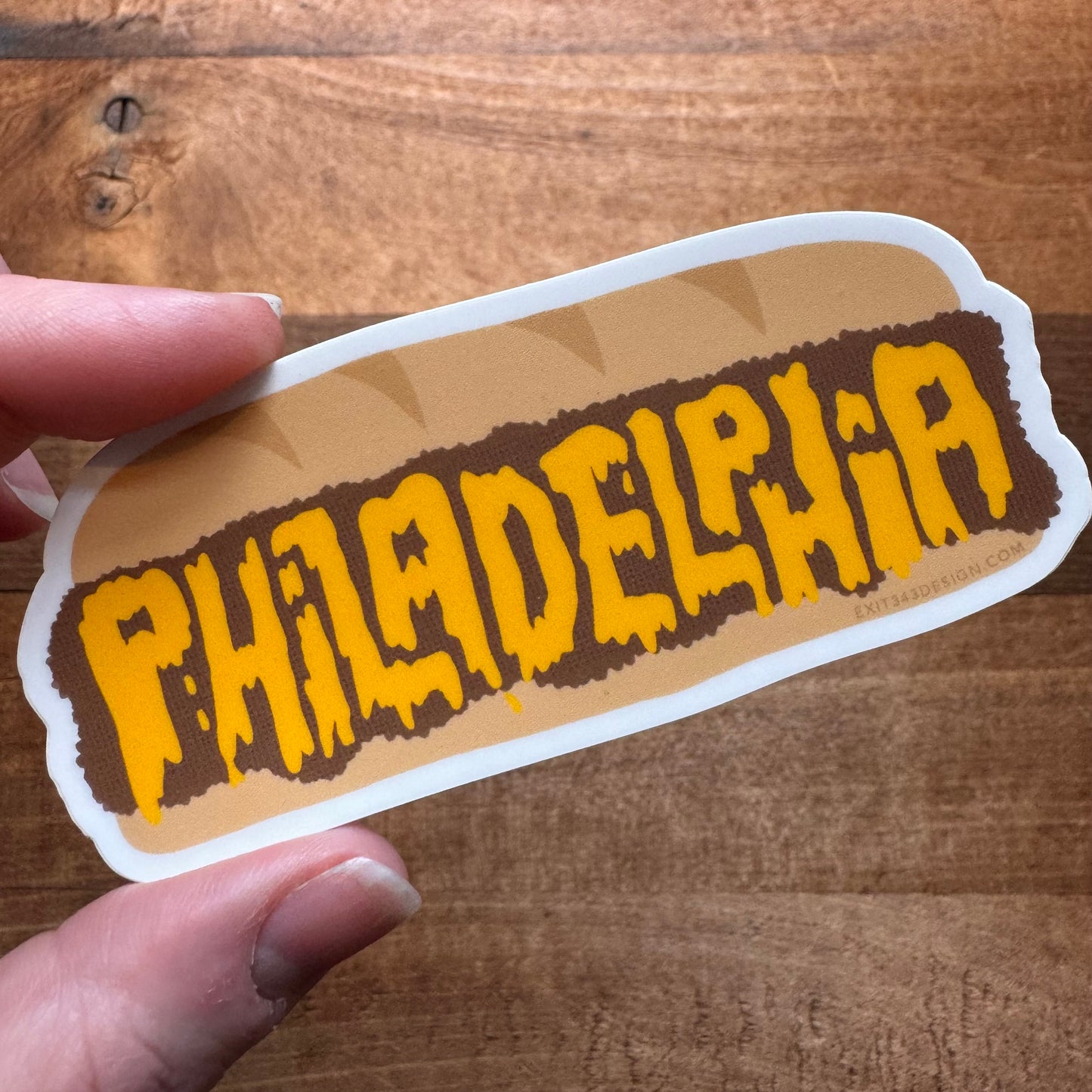 A hand holding a Philly Stickers with the word "Philadelphia" in a stylized font on a wooden surface, made by exit343design.