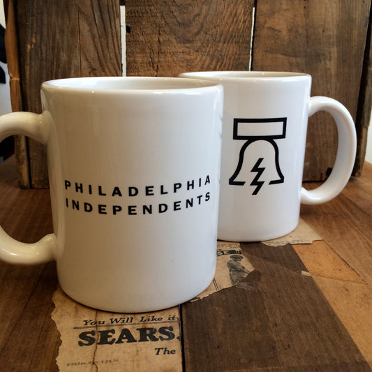 Two white coffee mugs displayed on a wooden surface: one Philadelphia Independents Bell & Bolt Mug with the text