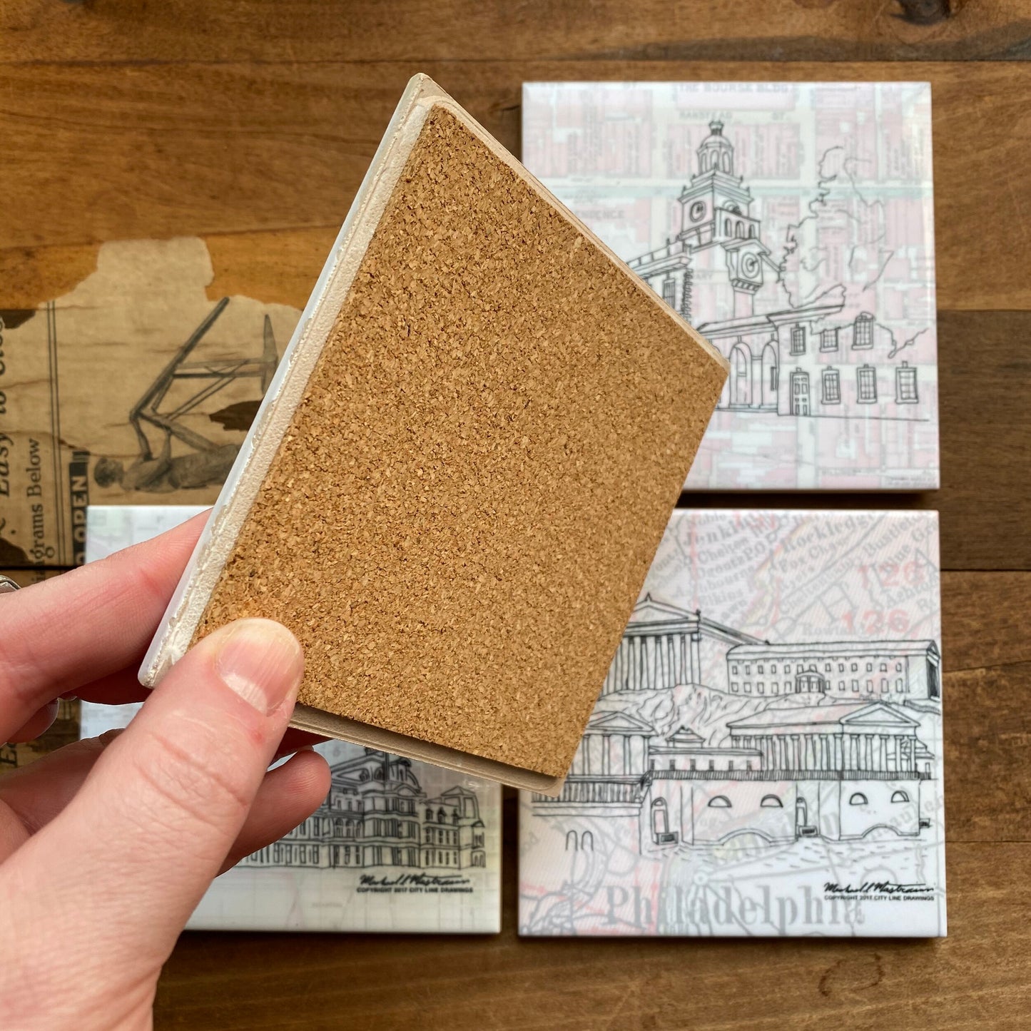 A person holding a Philly Landmark Coasters from City Line Drawings above a wooden table.