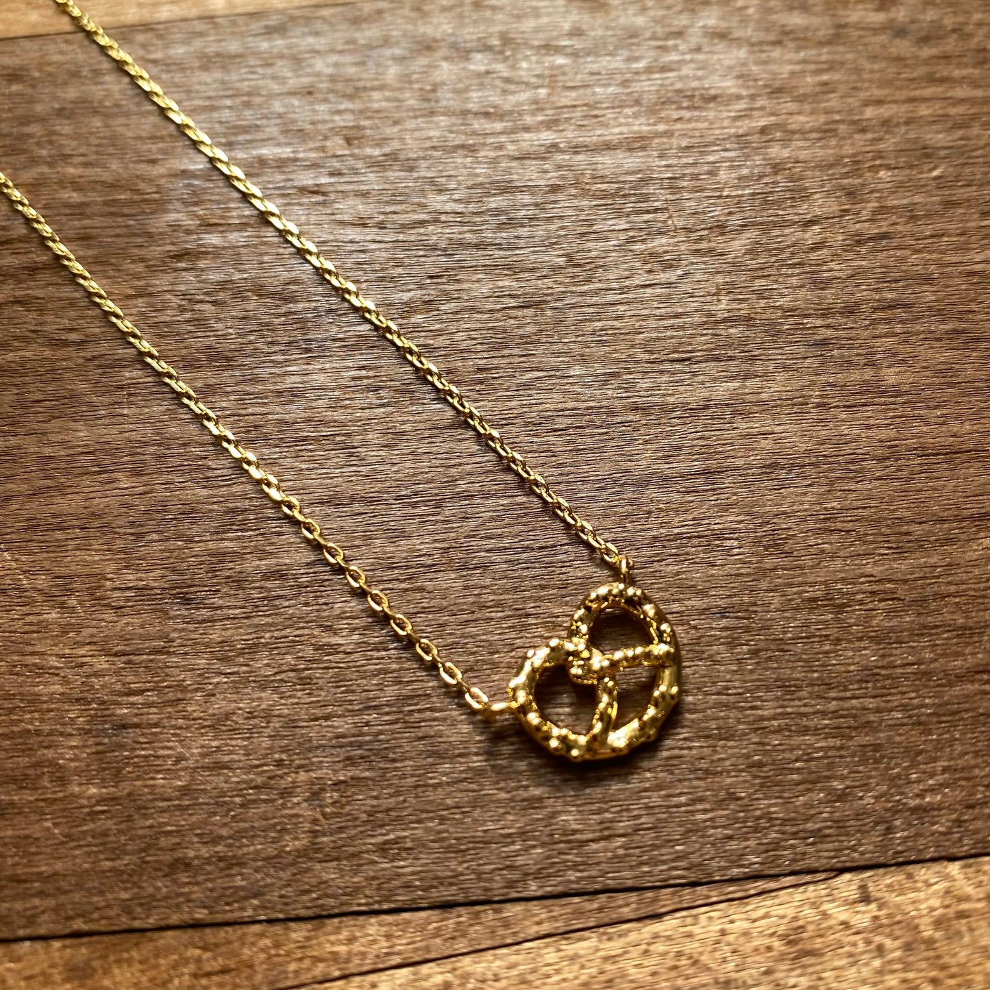 A gold Lily Lough LOVE & Pretzel necklace with a heart-shaped pendant on a wooden surface.
