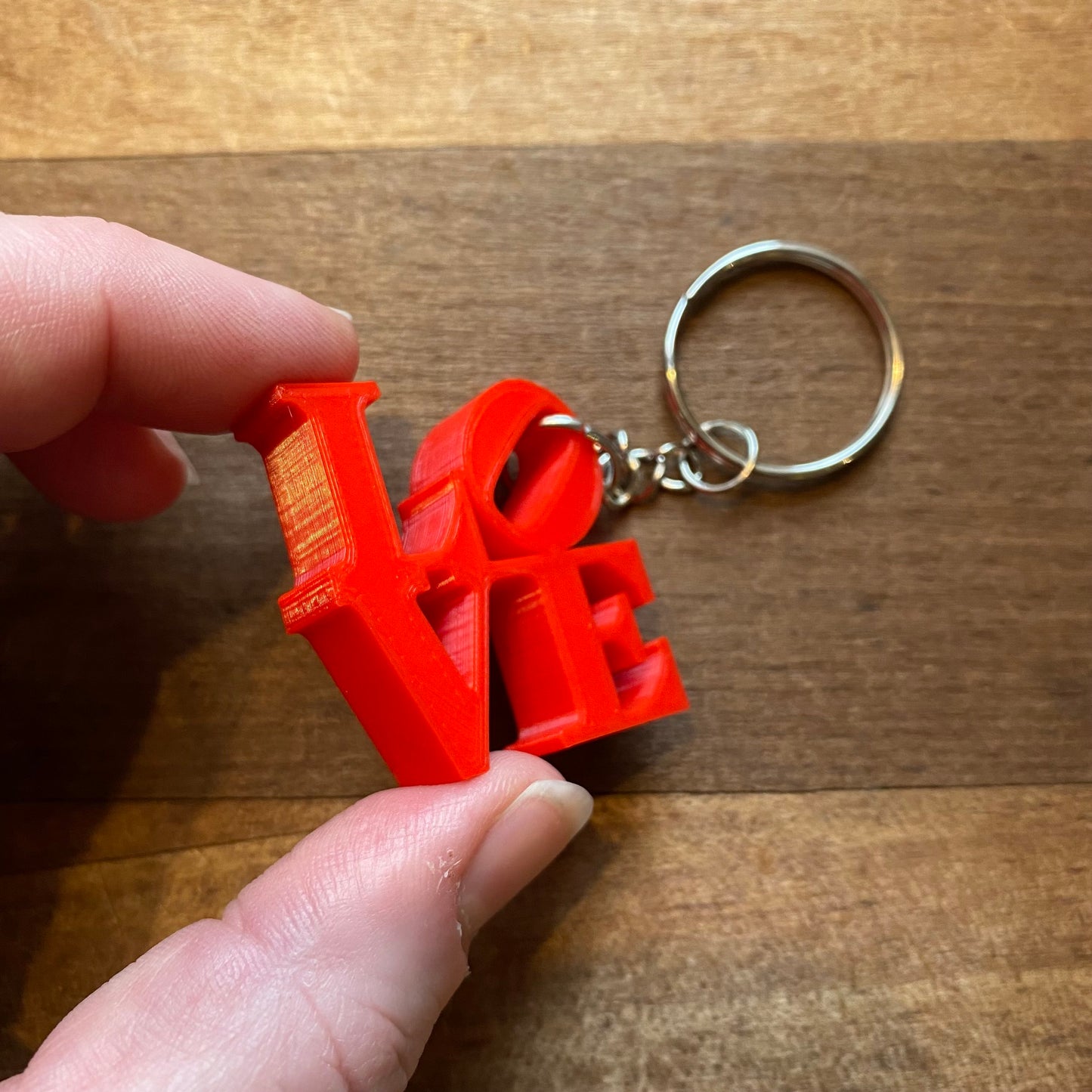 A hand holding a red "Rosewood Home LOVE" keychain with a wooden background.