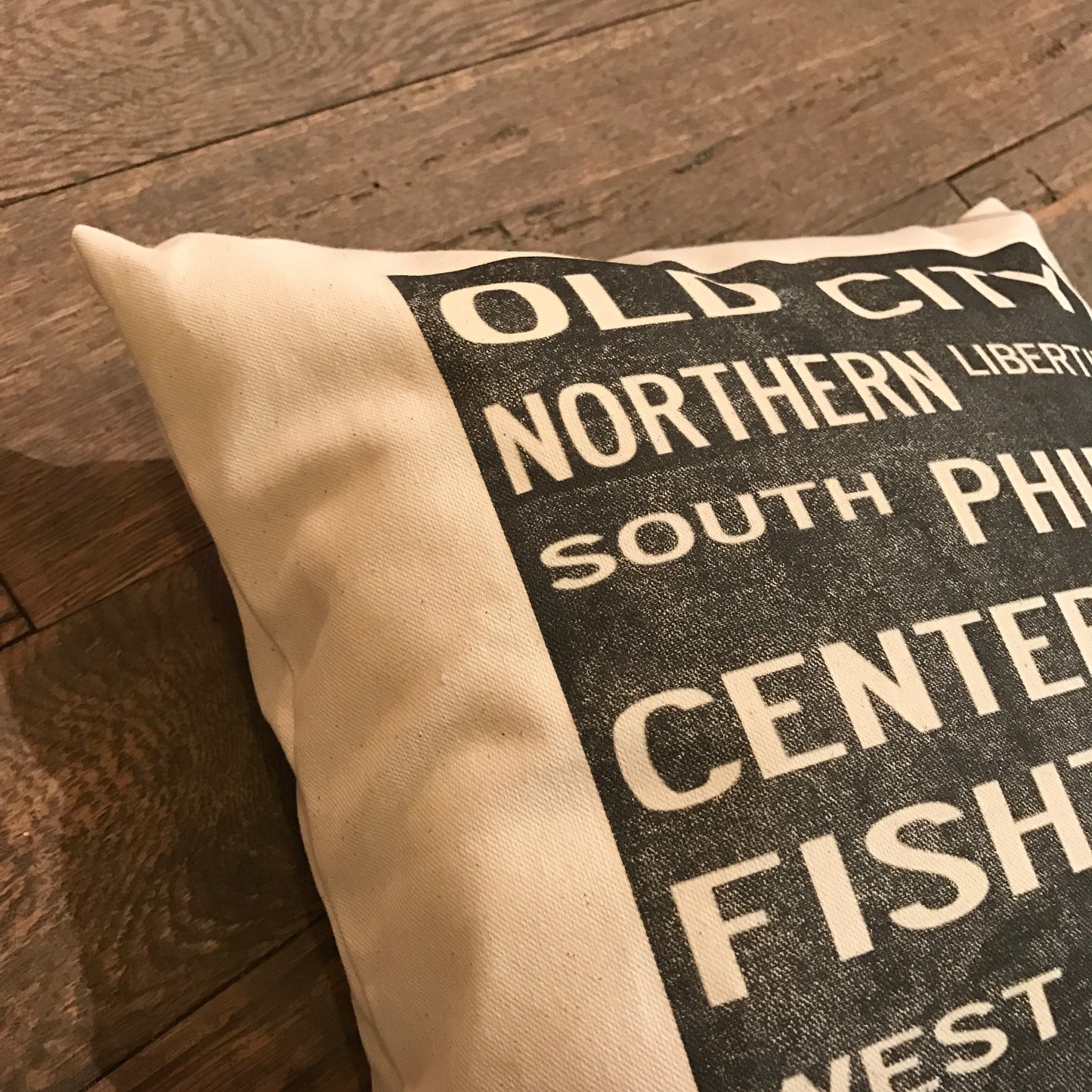 A decorative Philadelphia Neighborhoods Pillow from The Pillow Works with printed text "old city, northern liberties, south philadelphia, center city, fishtown, west philadelphia" on a wooden floor.