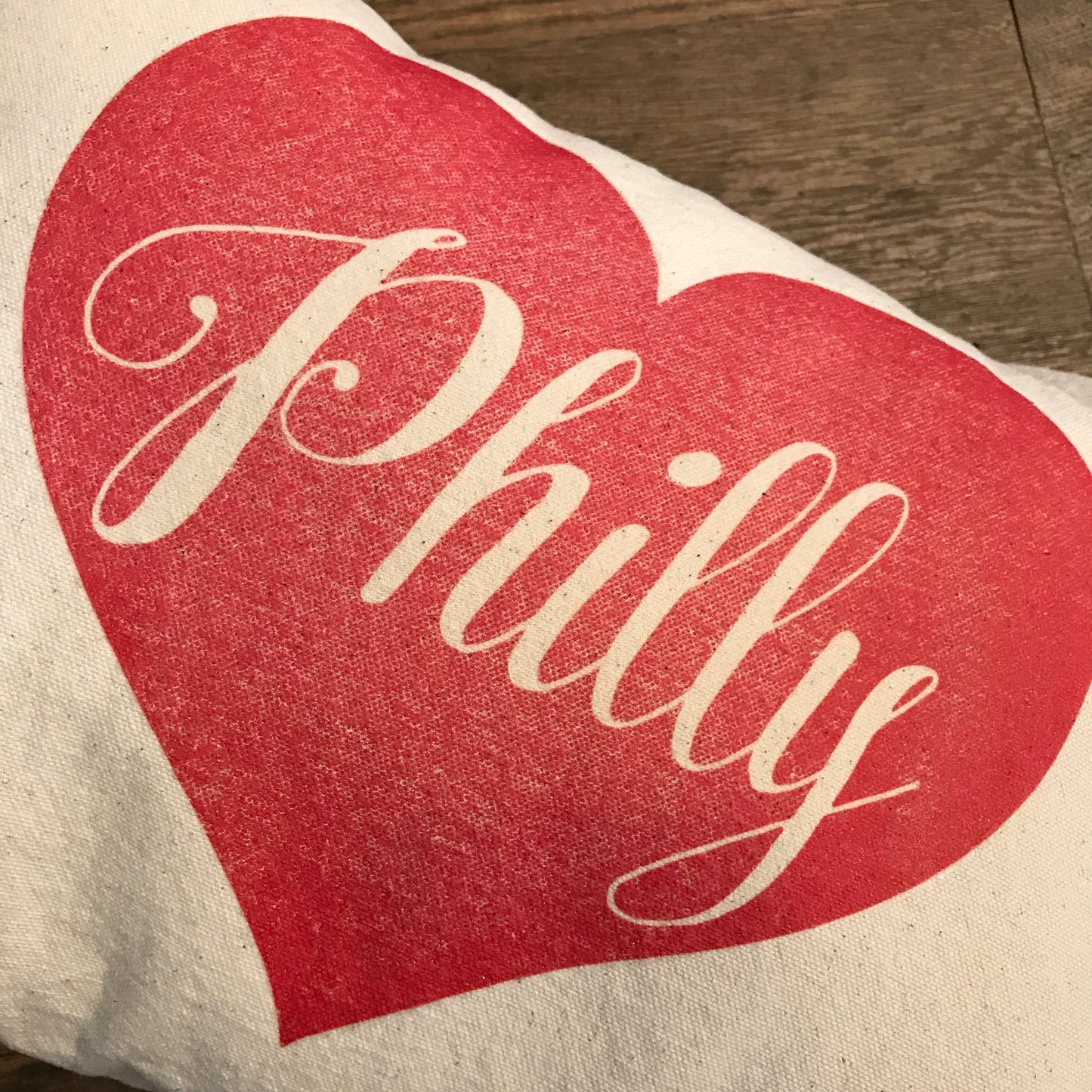 A red Philly Heart Pillow with the white cursive text "Philly" printed on an unbleached cotton drill fabric surface by The Pillow Works.