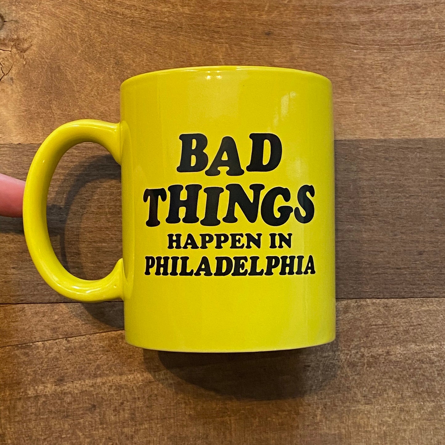 A yellow Bad Things Mug by Pink Bike Ralph with the text "bad things happen in Philadelphia" held by a person against a wooden background.
