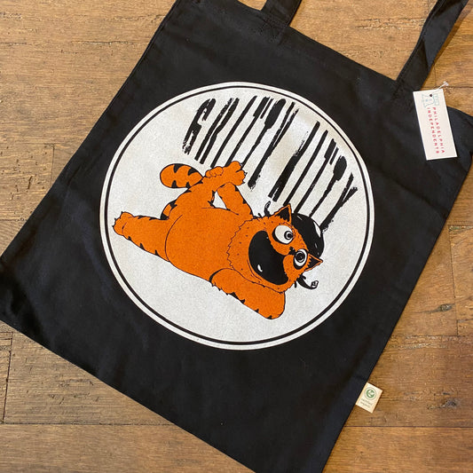 Gritty Kitty tote bag made from organic cotton with a circular design featuring a cartoon character resembling a furry, orange monster with big eyes, hanging from black bars by Slapstik.