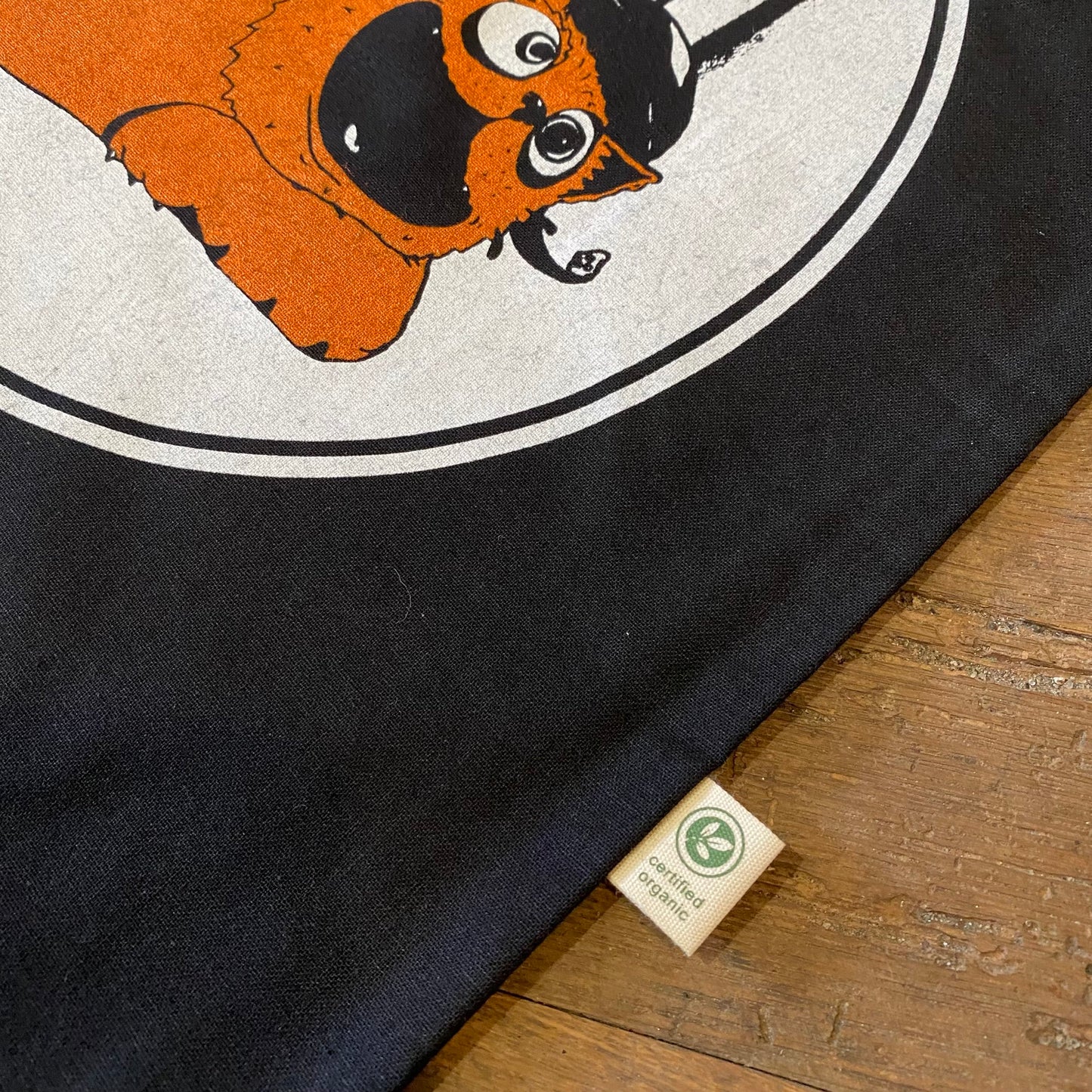 A Gritty Kitty Tote Bag with a circular white graphic featuring an orange cartoon character, lying on a wooden surface with a visible size tag, made from organic cotton. Manufactured by Slapstik.