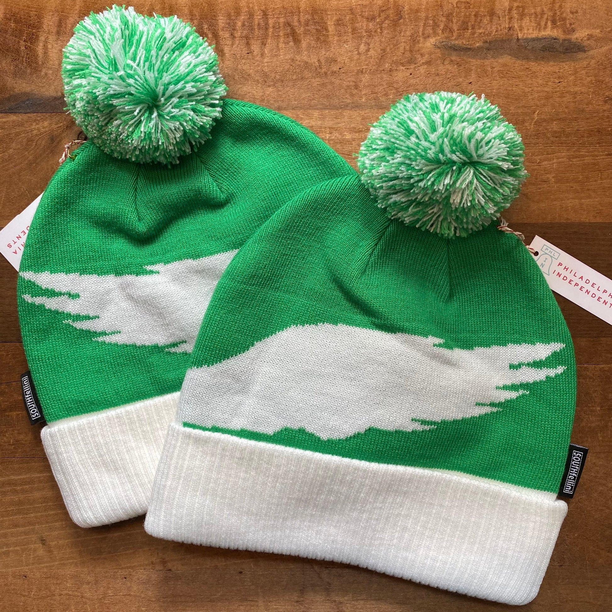 Two green-and-white, 100% polyester knit Helmet Head Eagles Beanies by South Fellini with pom-poms and white wing designs are displayed on a wooden surface. White tags are attached to each one size fits all beanie.