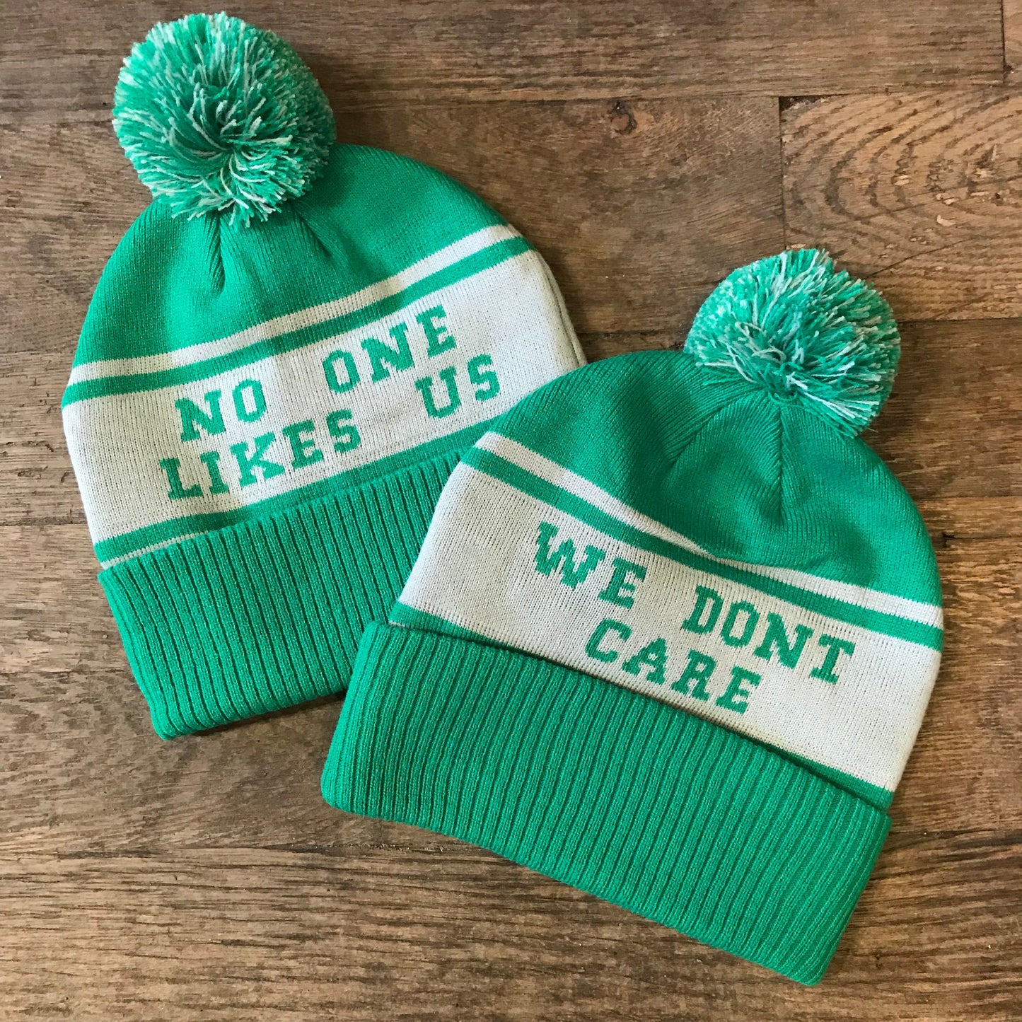 Two South Fellini green and white knit beanies with pom-poms sit on a wooden surface. One beanie reads "NO ONE LIKES US" and the other reads "WE DON'T CARE." Perfect for Philly fans, these hats capture the spirit of the Philadelphia Eagles' Super Bowl Parade.
