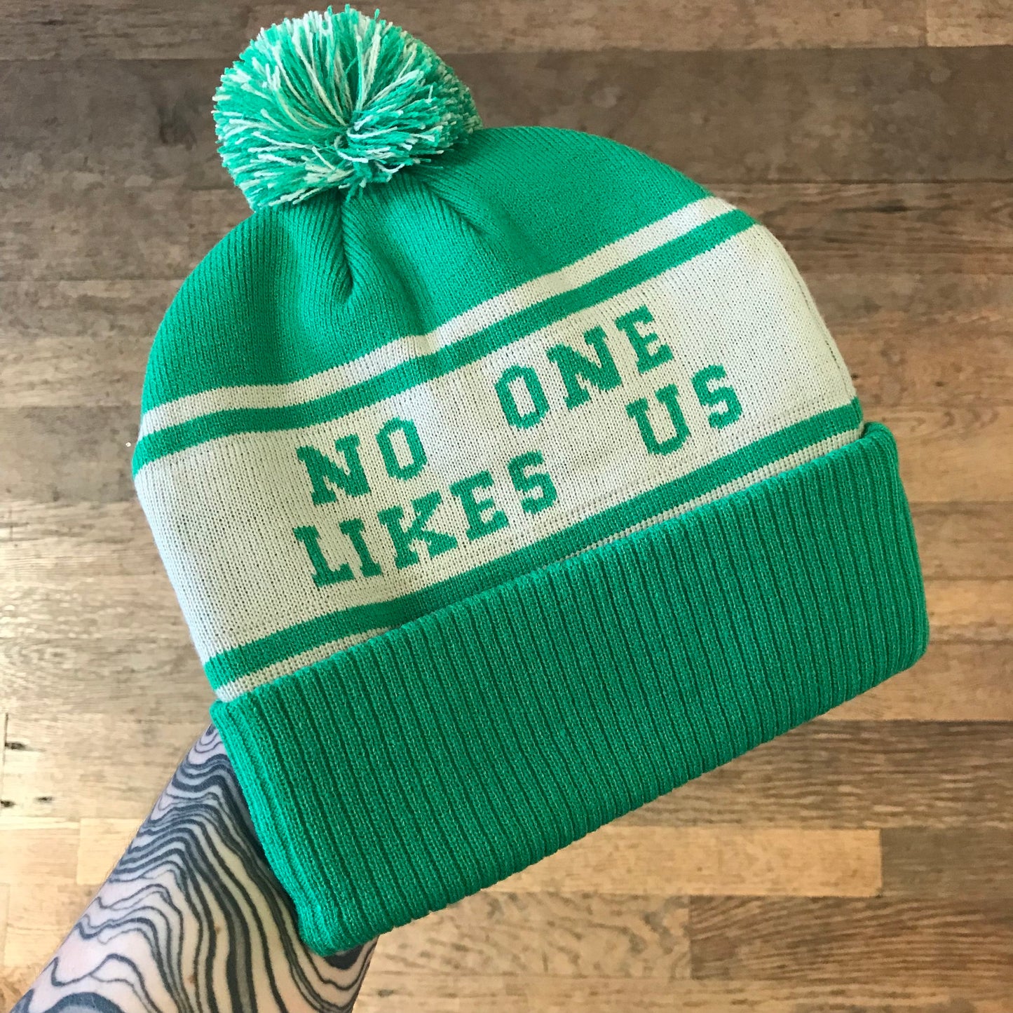 A tattooed hand holds the South Fellini "No One Likes Us Eagles Beanie I," a green and white beanie adorned with a pom-pom and the spirited phrase "NO ONE LIKES US," capturing the vibrant essence of Philly as if ready for a Super Bowl Parade celebrating the Philadelphia Eagles.