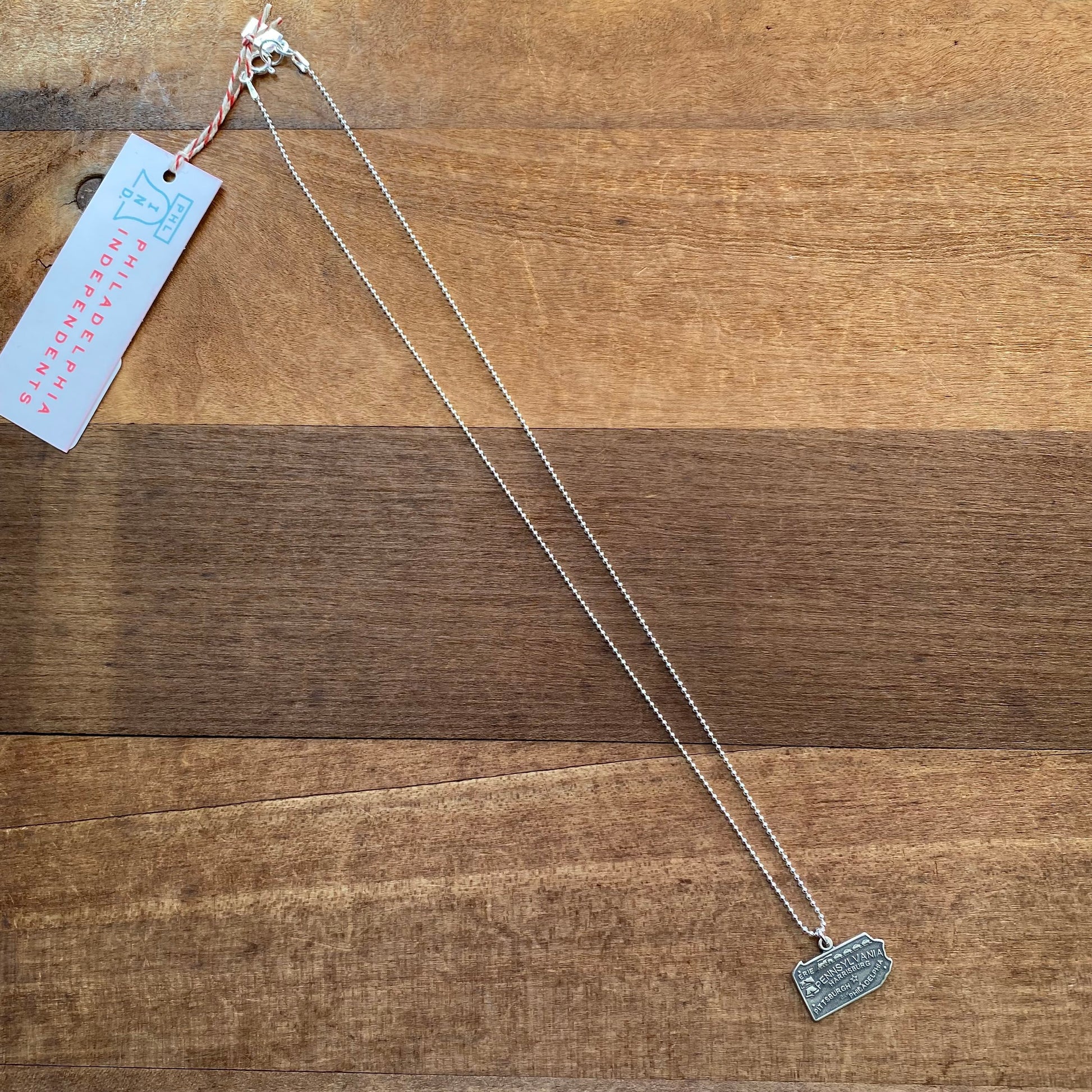 The Pennsylvania Necklace by Stone Cooper, a sterling silver piece featuring a small rectangular pendant reminiscent of the Pennsylvania state shape, is displayed on a brown wooden surface. A pink hang tag reading "Stone Cooper" is attached to the necklace and visible on the left side.