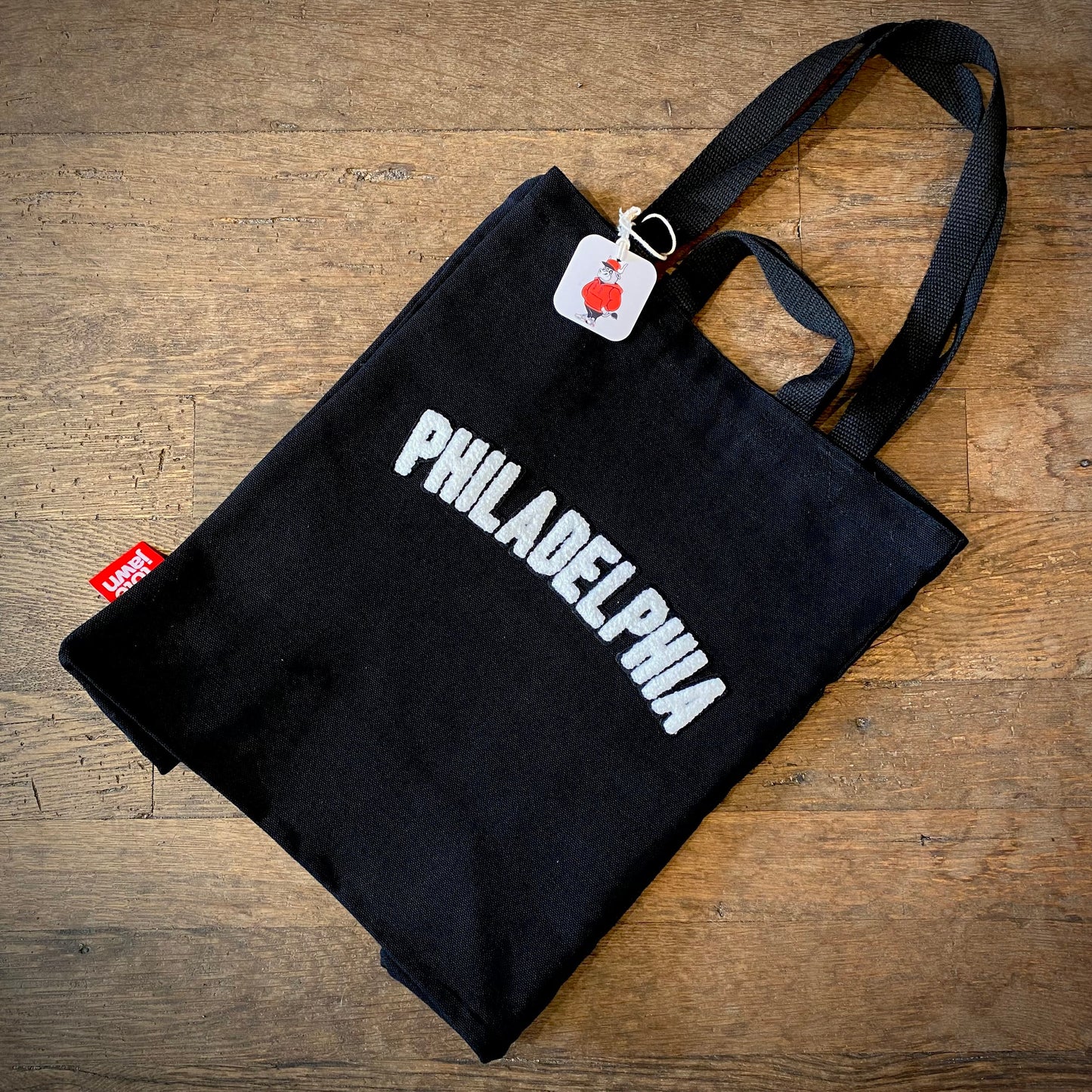 Black canvas Philadelphia Patch Tote Bag with the word "Philadelphia" in white letters, featuring a red Tote Jawn logo tag and placed on a wooden surface.