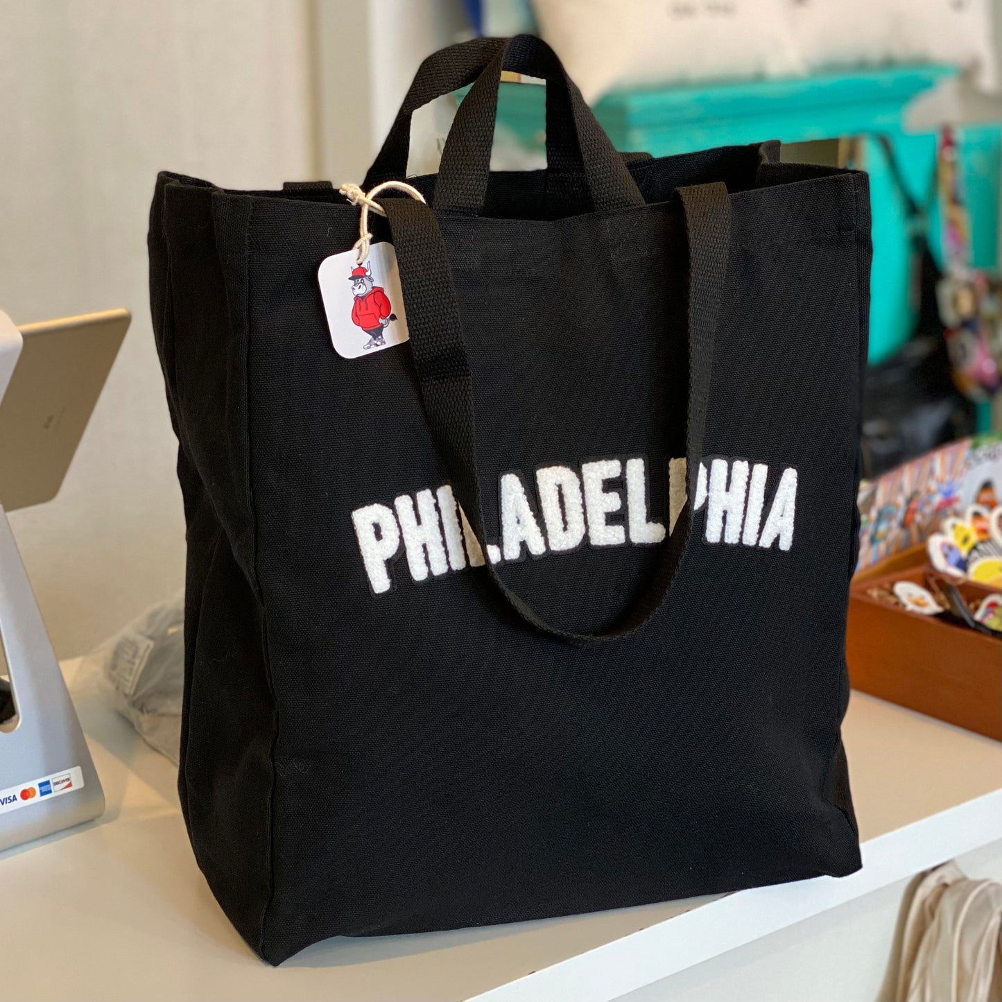 Black canvas Philadelphia Patch tote bag with the word "Philadelphia" in white letters on a tabletop, featuring a small tag shaped like a basketball jersey by Tote Jawn.