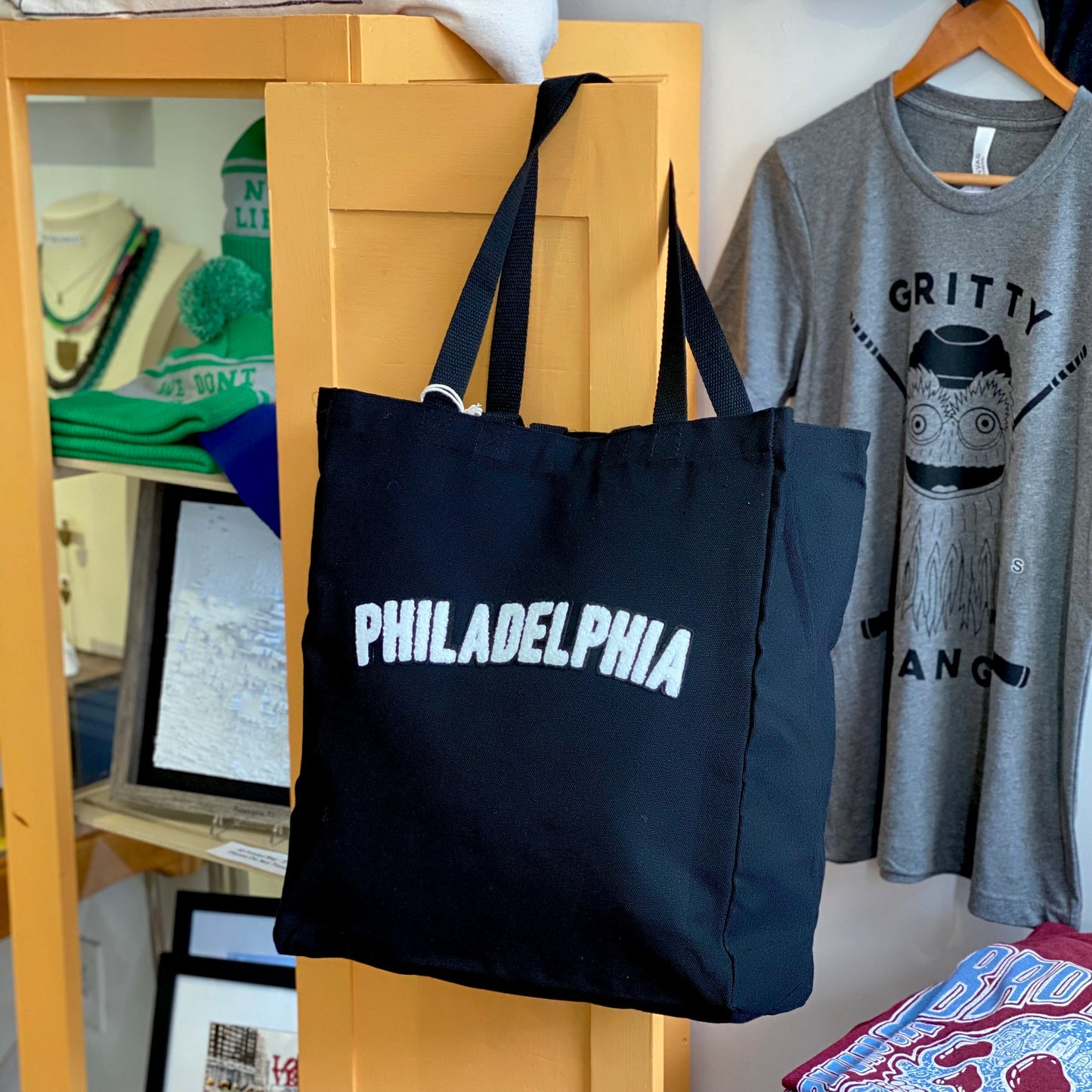 Black canvas Philadelphia Patch Tote Bag with "Philadelphia" printed in white, hanging on a yellow wooden stand in a store with assorted merchandise in the background.