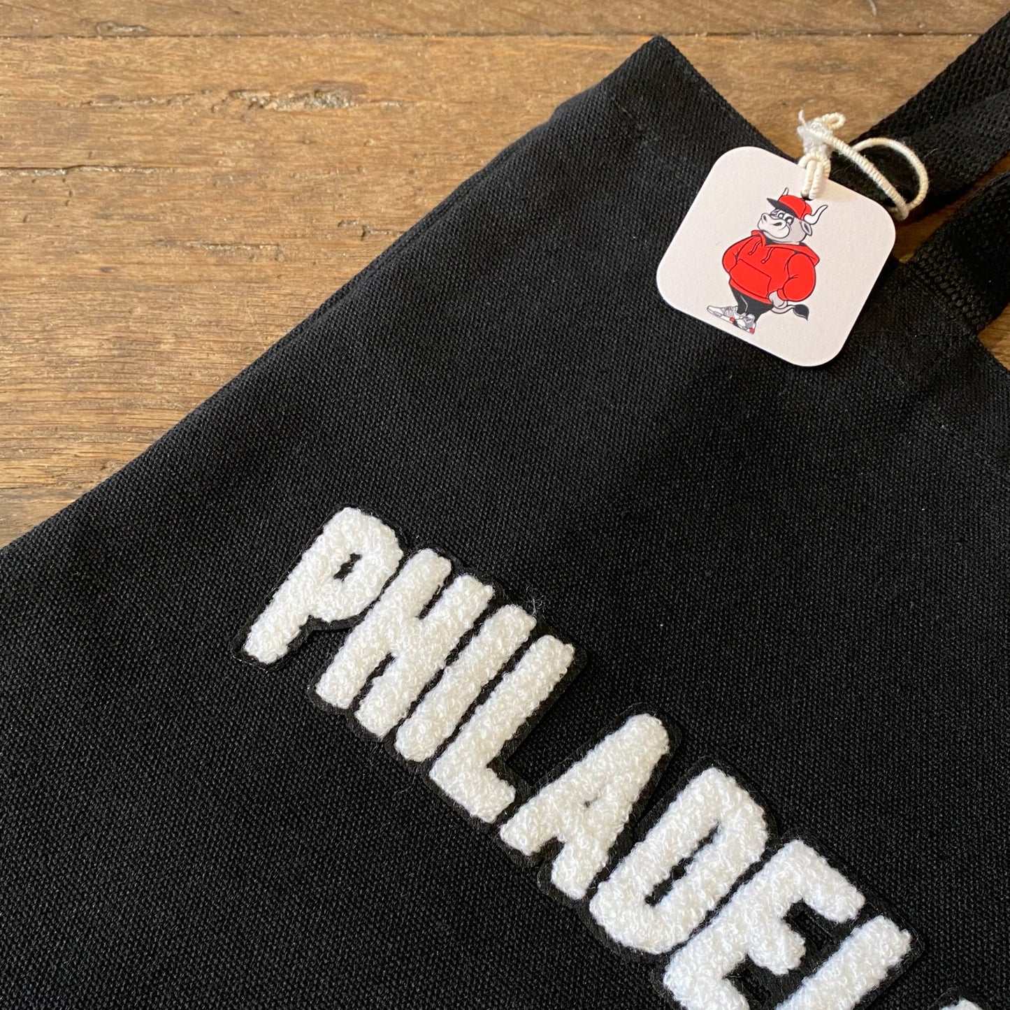 Black canvas Philadelphia Patch Tote Bag with the word "Philadelphia" in white letters, featuring a tag with a cartoon of a muscular chef by Tote Jawn.