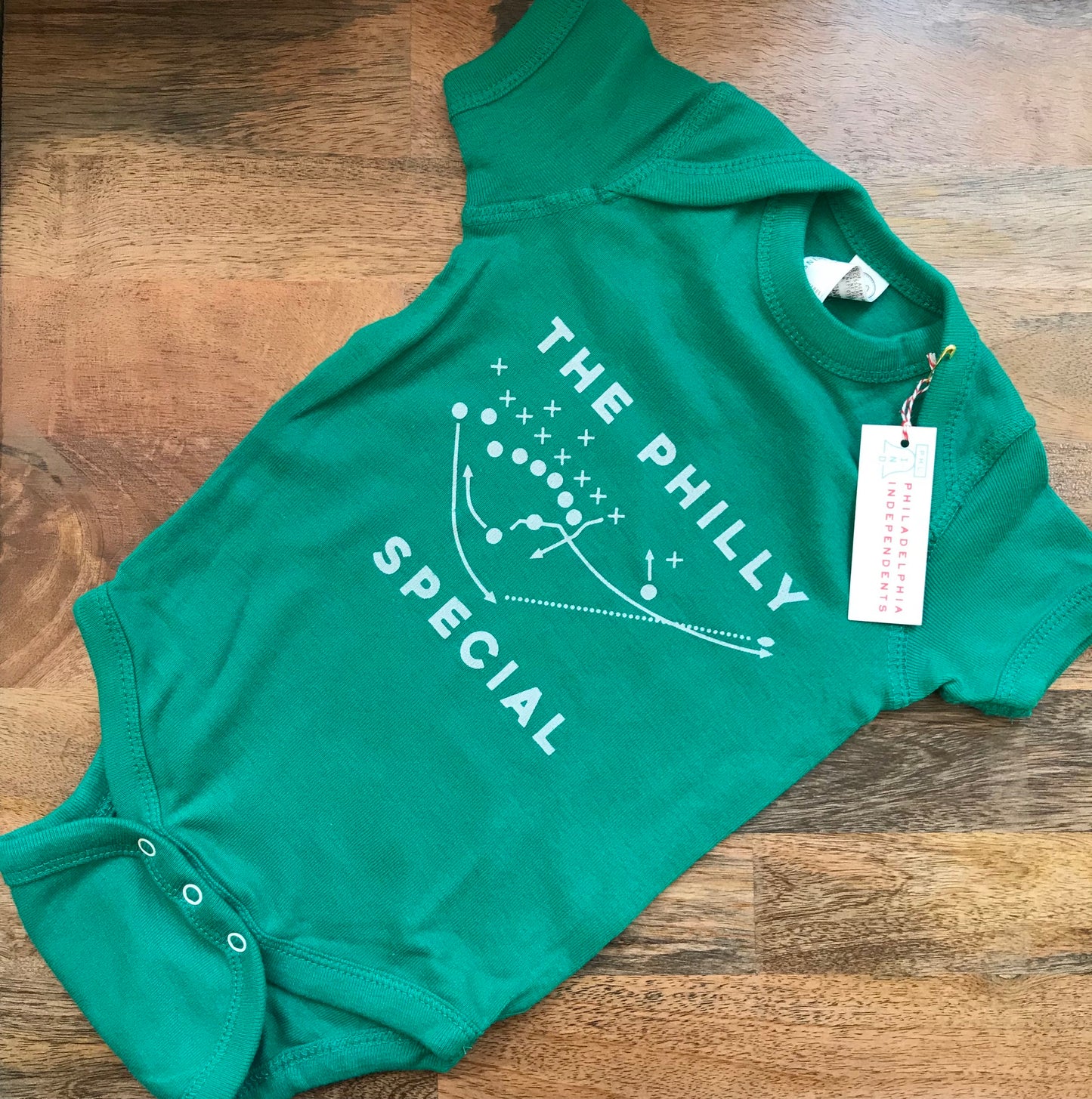 Green exit343design Philly Special Baby Onesie with "the philly special" play diagram and a price tag on a wooden floor.