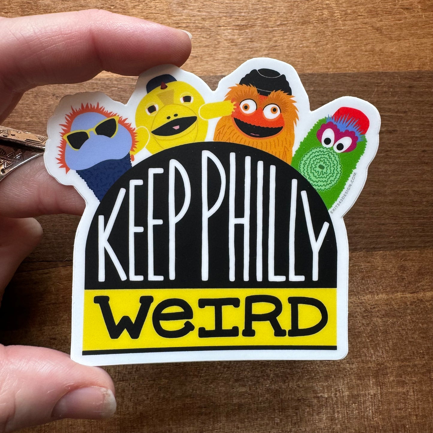 A hand is holding a Philly-themed vinyl sticker from exit343design, featuring colorful, cartoonish characters including Gritty, above the text "KEEP PHILLY WEIRD" on a wooden background.